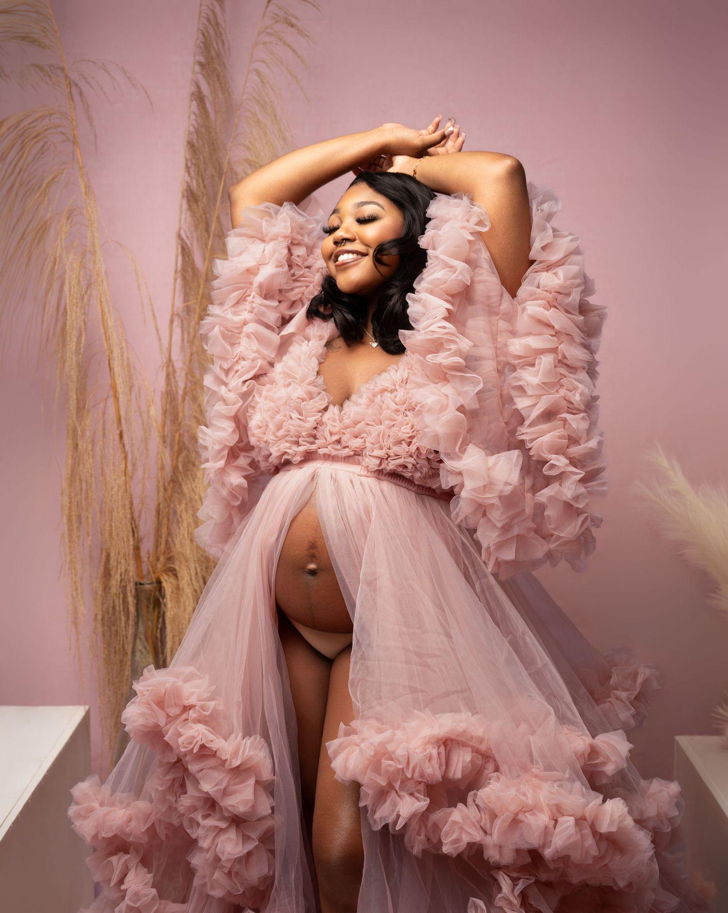 Pregnant woman posing in a pink ruffled tulle maternity robe, showcasing her baby bump in an elegant studio shoot.