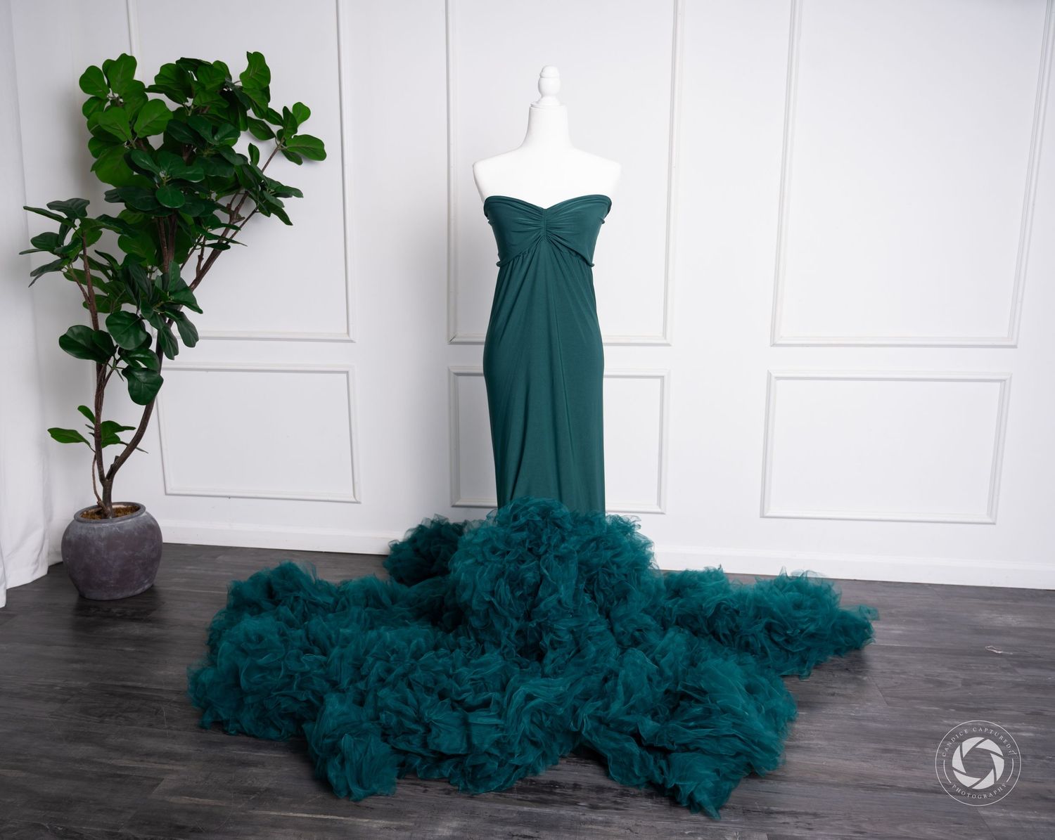 A teal mermaid-style evening gown with dramatic ruffled tulle train on a light colored floor.