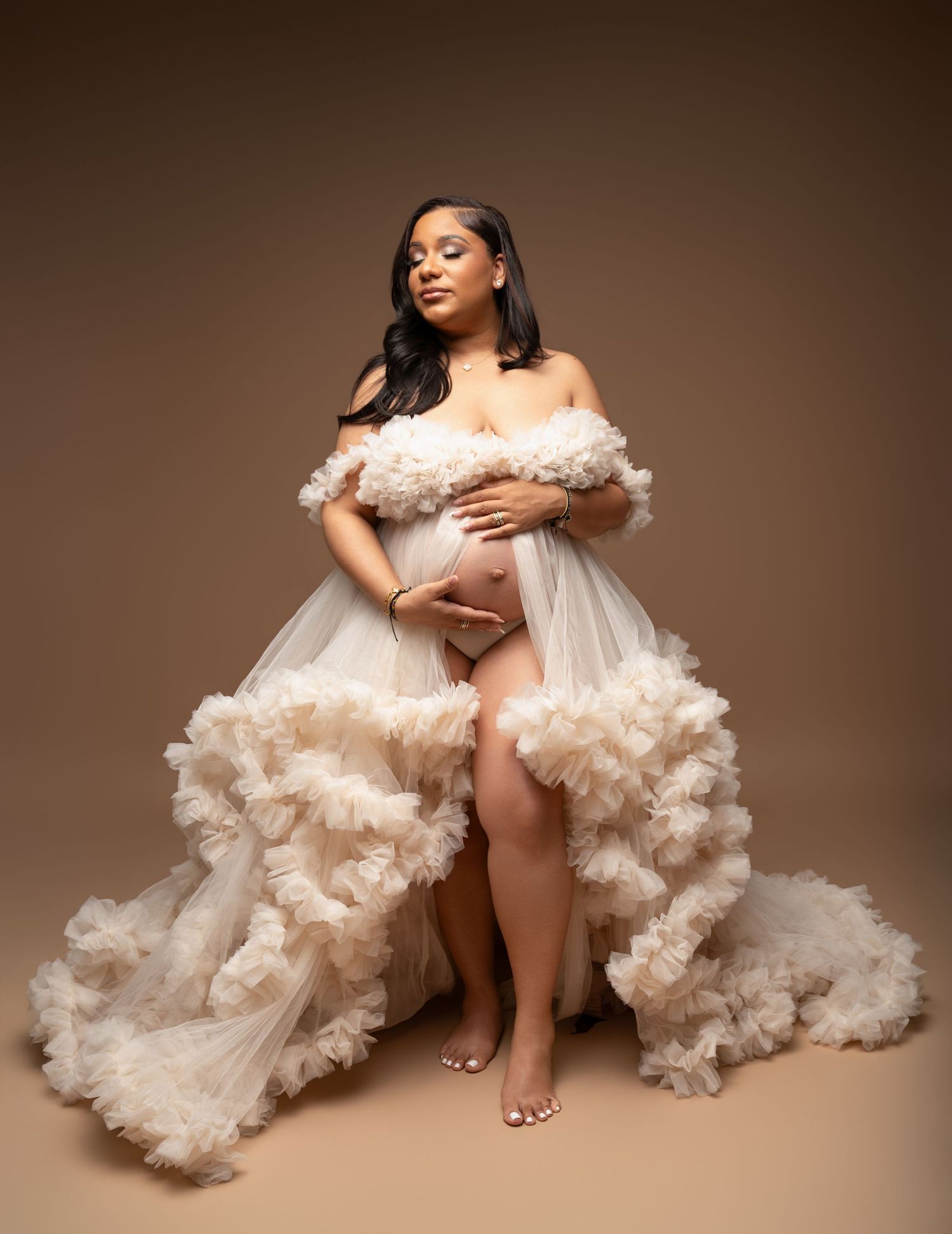 Pregnant woman in ruffled off-shoulder cream tulle maternity gown posing gracefully in a professional studio photo.