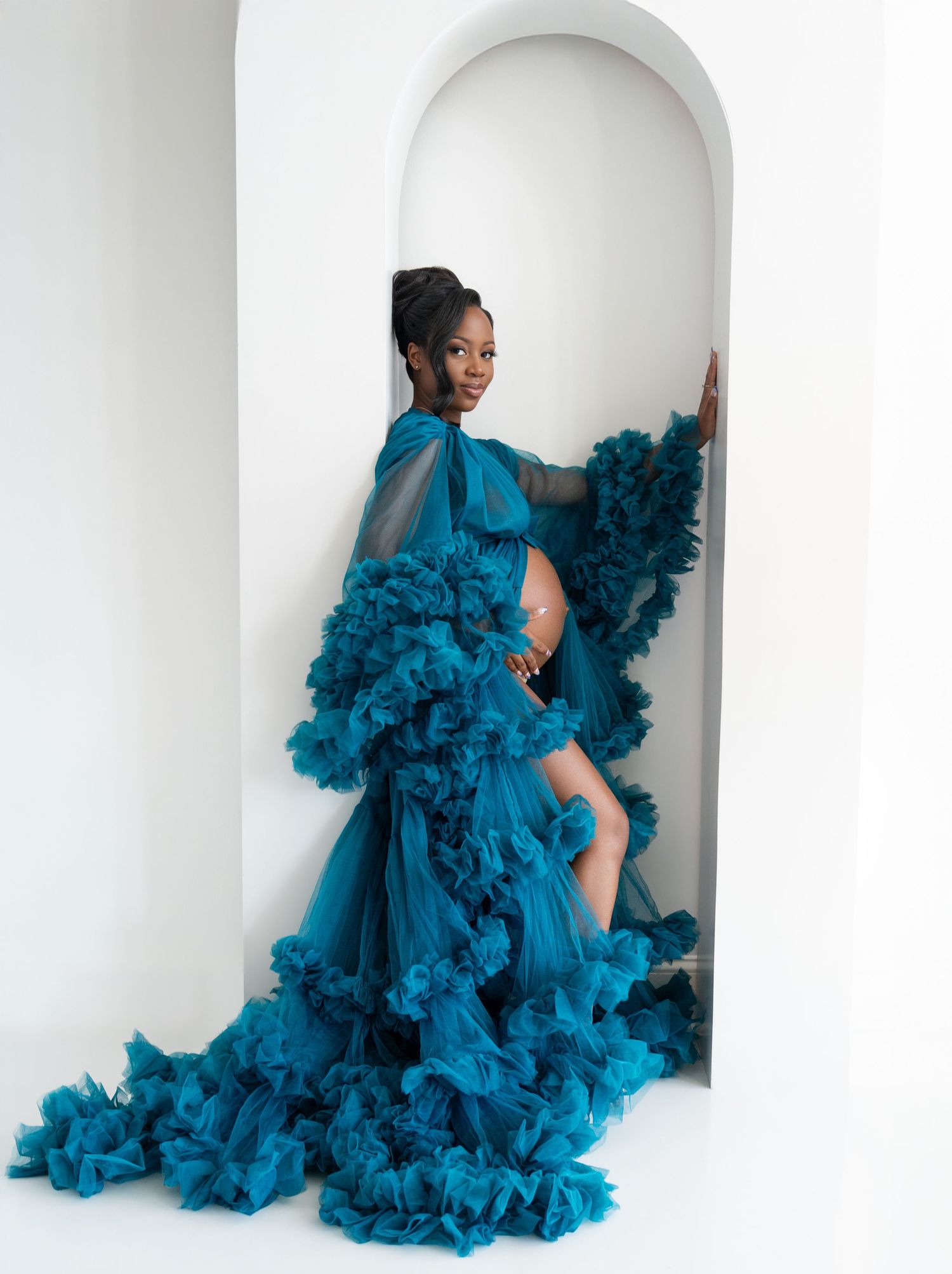 Pregnant woman in a stunning teal ruffled tulle maternity gown posing elegantly in a white arched doorway.