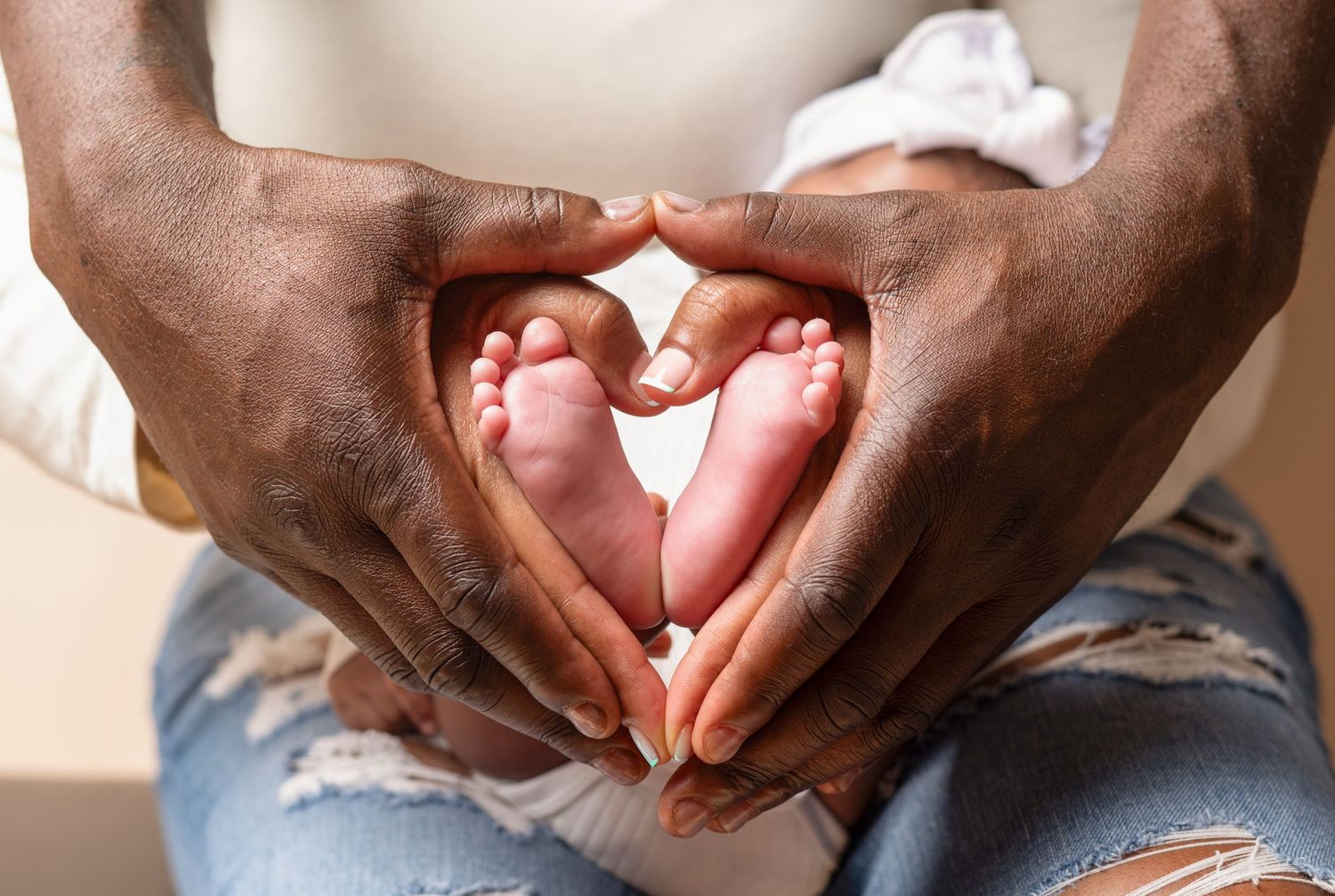 Dark hands form a heart shape around tiny baby feet captured in tender moment.