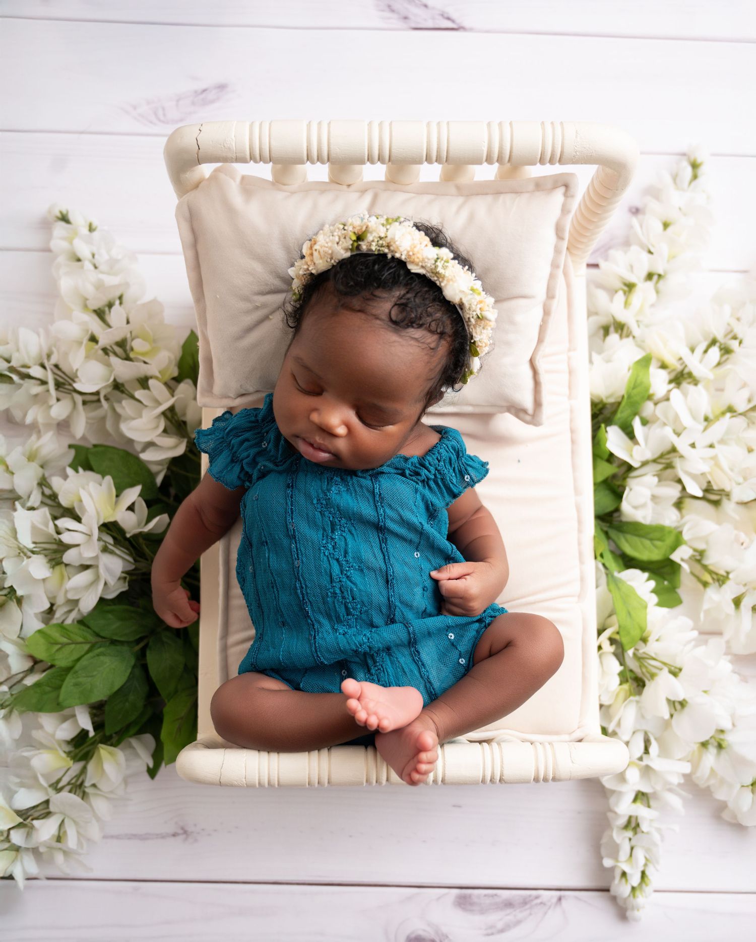 Newborn baby girl in teal romper and floral headband sleeping peacefully on a tiny white bed surrounded by white flowers.