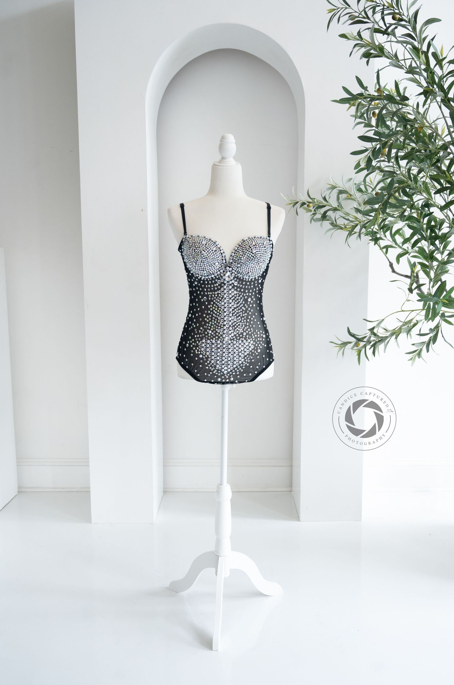 A black lace corset bustier displayed on white mannequin stand in front of arched alcove with decorative plant.