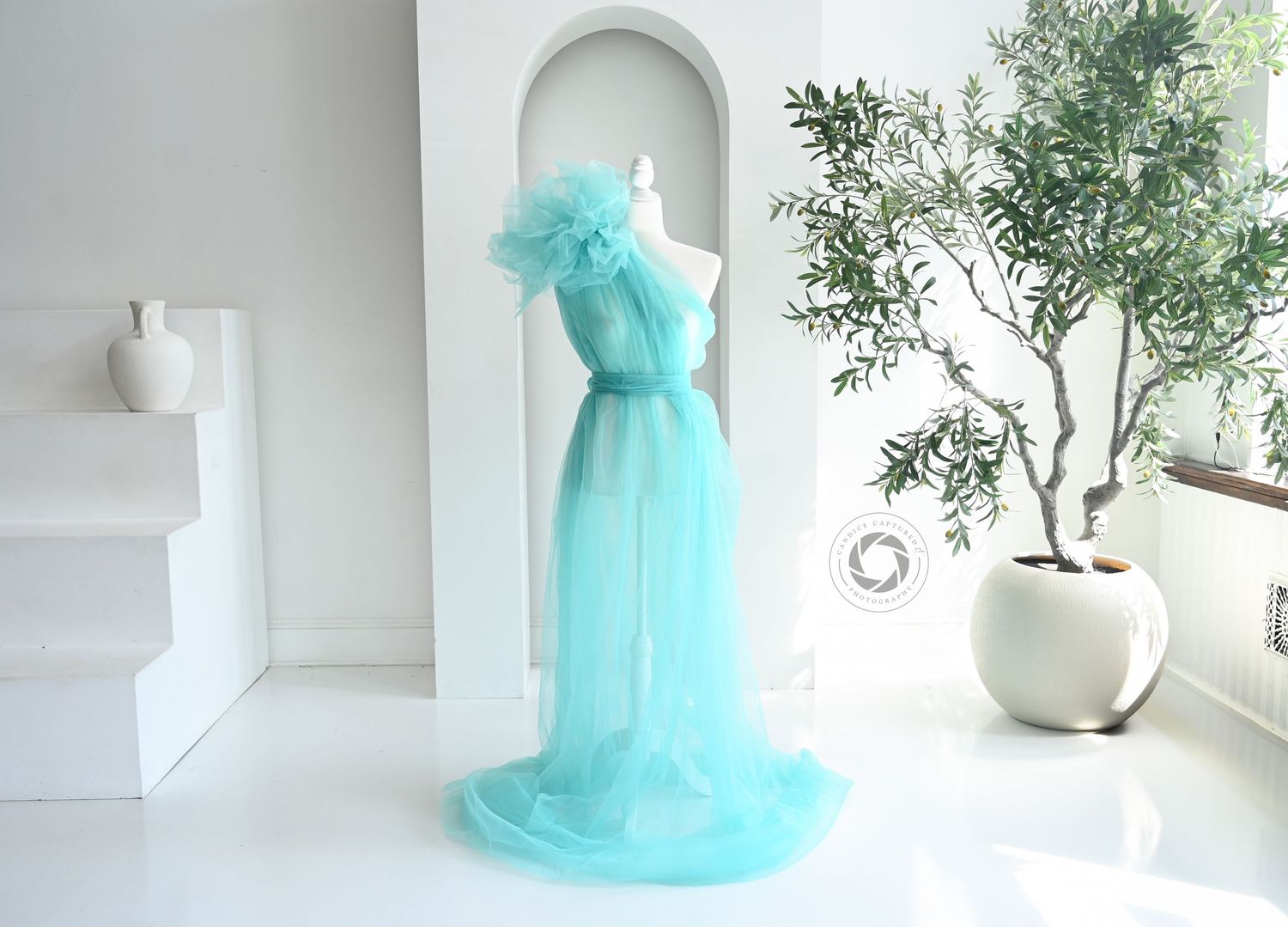A bright teal tulle ballgown dress on a mannequin photographed against a white wall with natural window light.