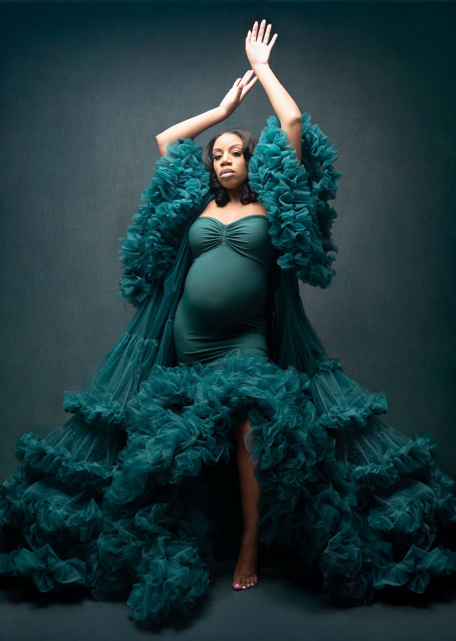 Maternity photoshoot featuring a dramatic teal ruffled tulle robe against a dark background.