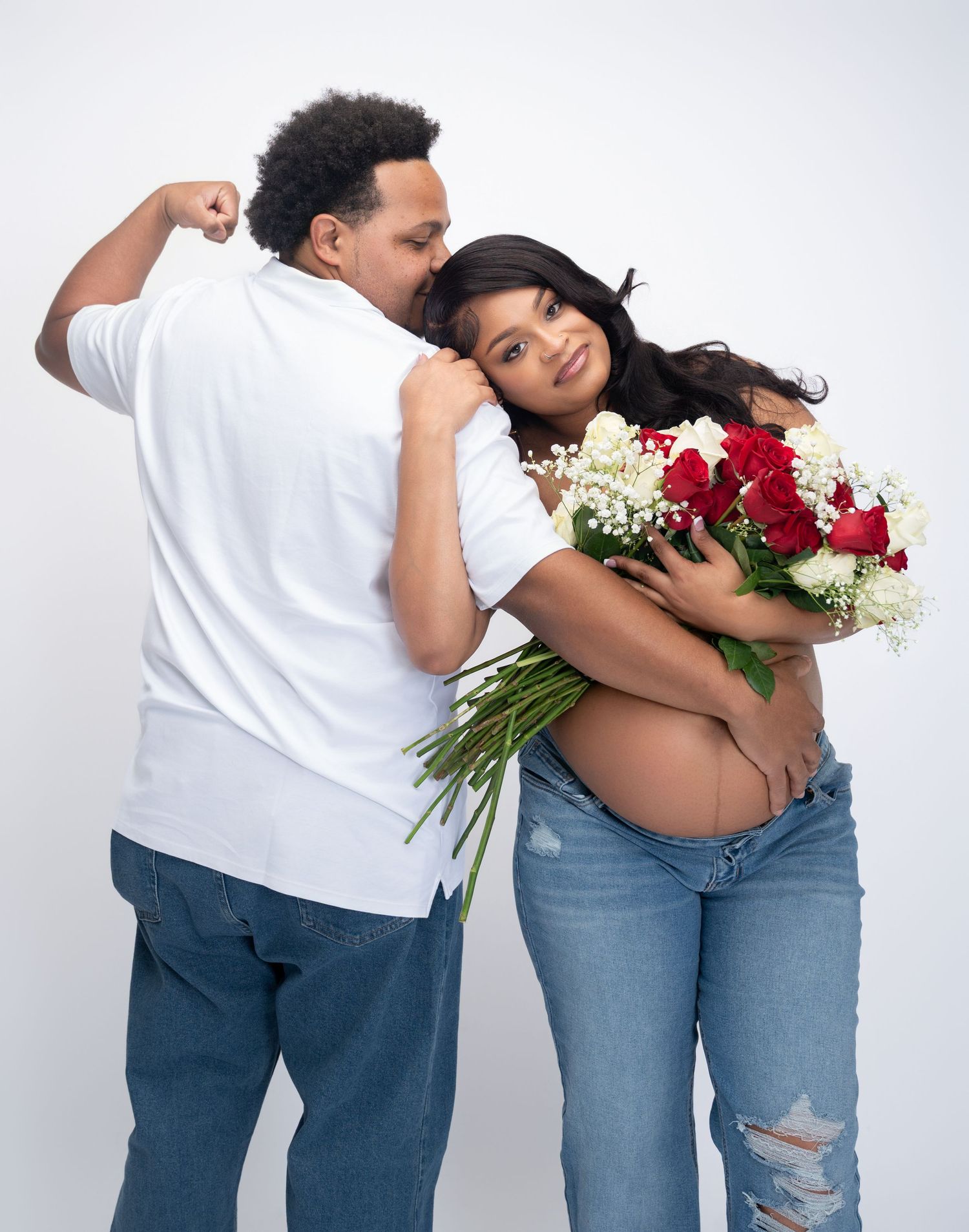 Pregnant woman holding red and white roses embraced by partner in white shirt and jeans on white background.