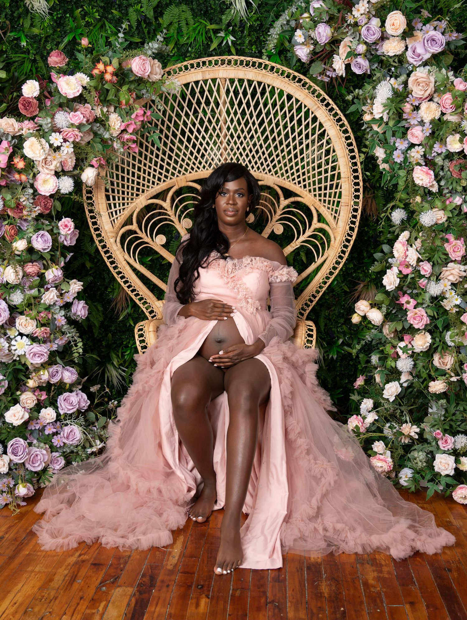 A decorative peacock chair surrounded by pink and white roses with a flowing pink tulle gown creating an ethereal maternity scene.