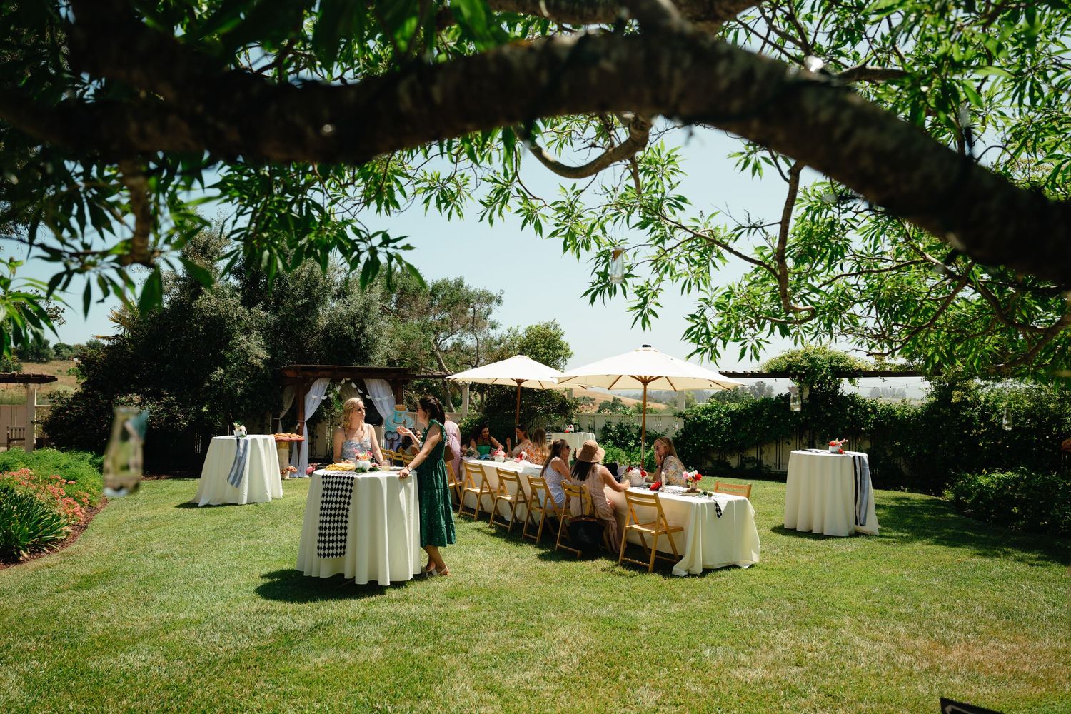Outdoor garden wedding reception with long dining table, white cocktail tables, and umbrellas on lush green lawn.