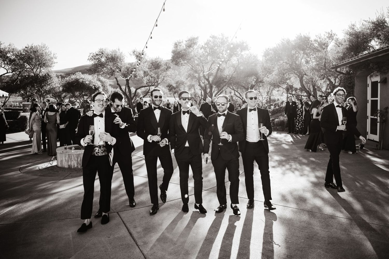 Groomsmen in black tuxedos walk confidently outdoors in a black and white wedding photo.