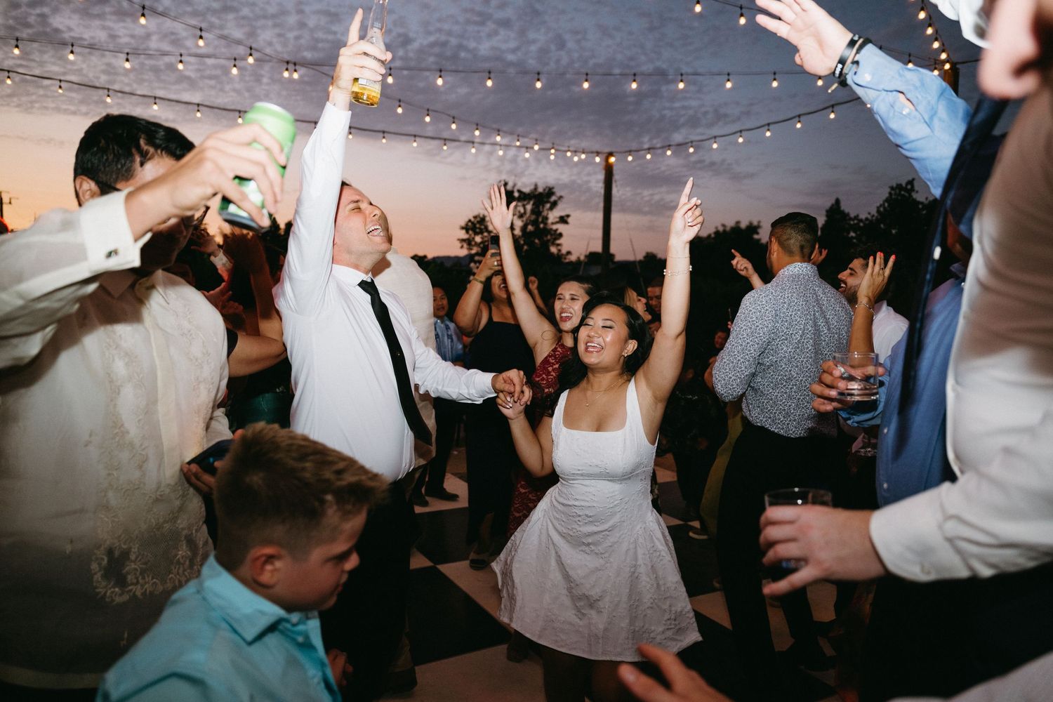 Wedding reception party guests dance and celebrate on a crowded dance floor under string lights and decorations.