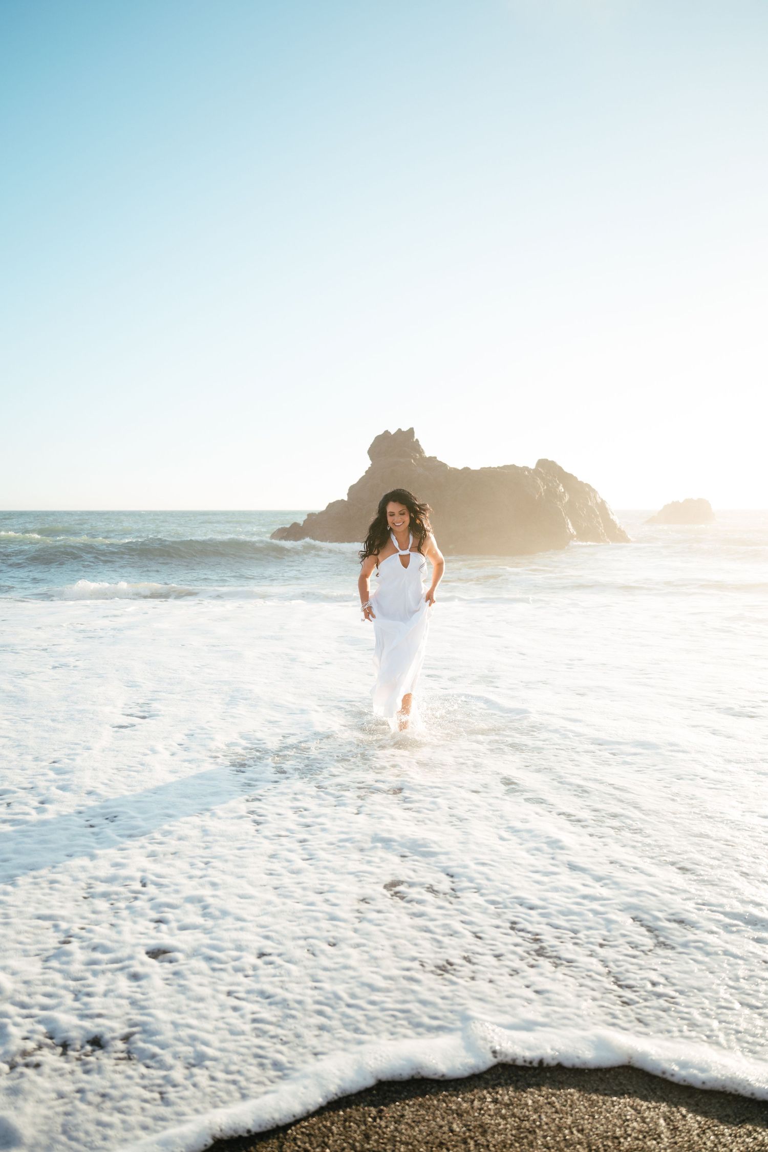 A figure in a flowing white dress walks along a misty beach shoreline at sunset with waves crashing nearby.