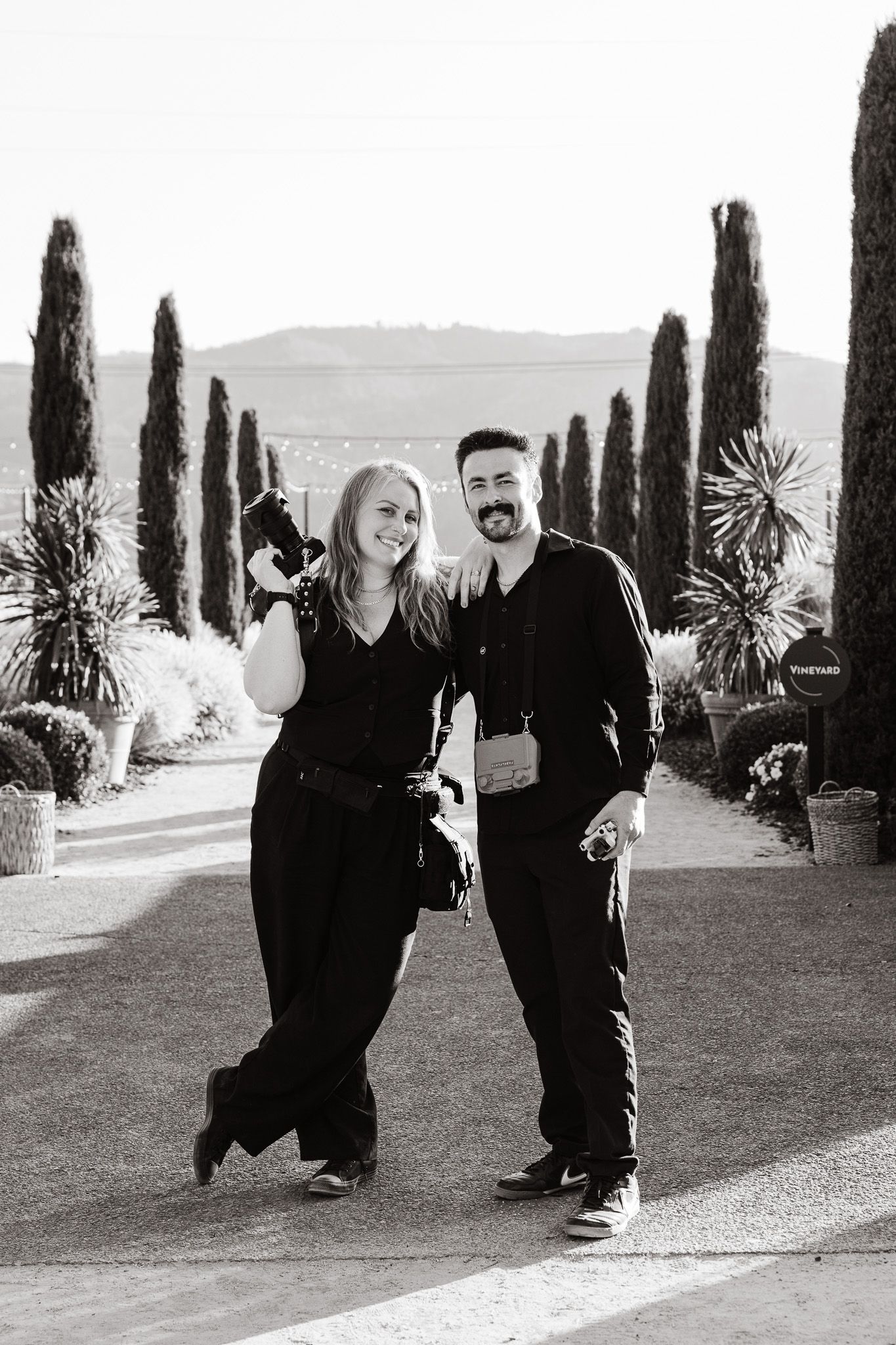 Black and white photo of two photographers smiling and holding cameras outdoors with cypress trees and mountains behind them.