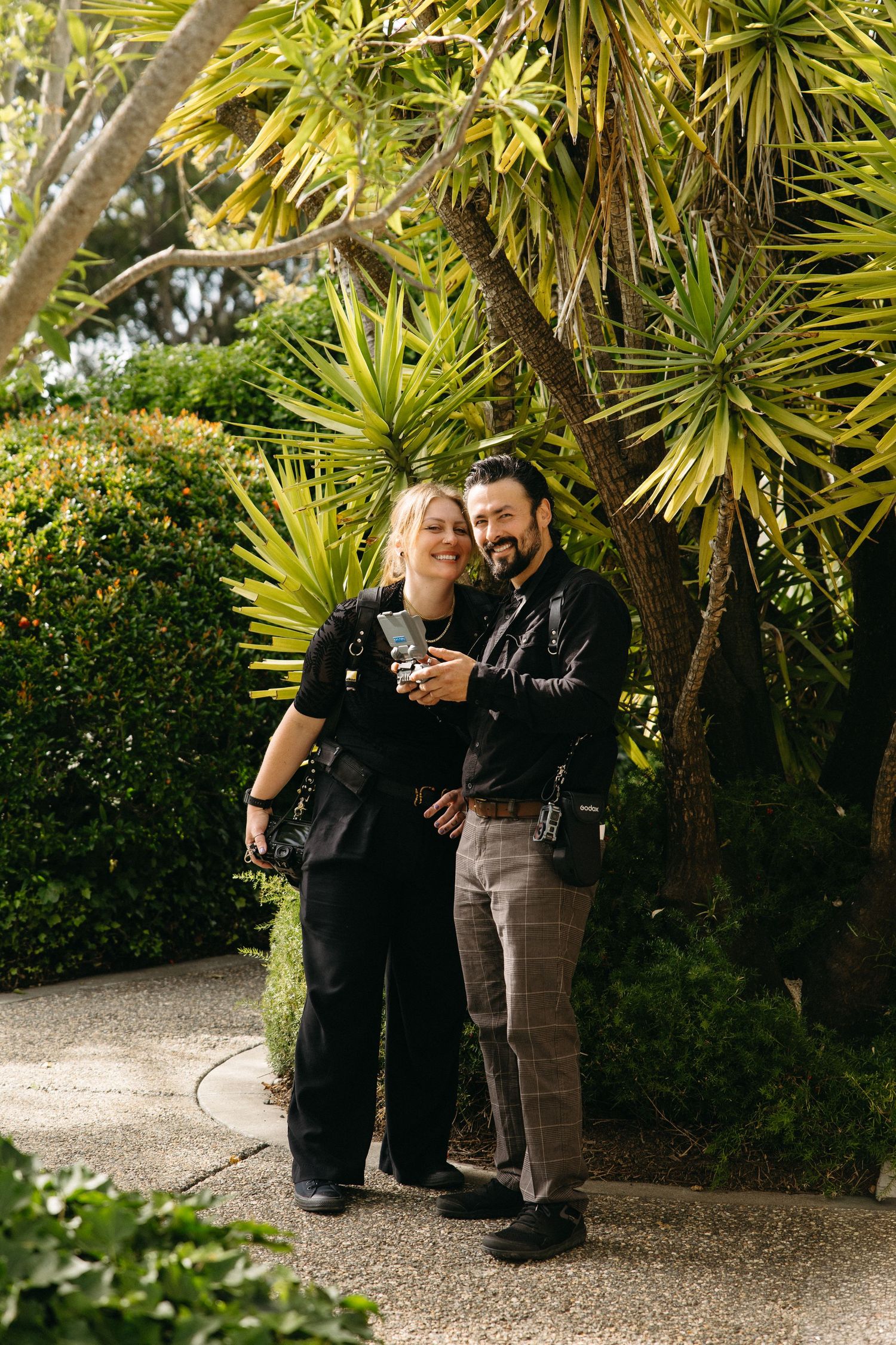 A smiling couple poses together in a lush garden, dressed in black outfits, holding cameras among tropical plants.