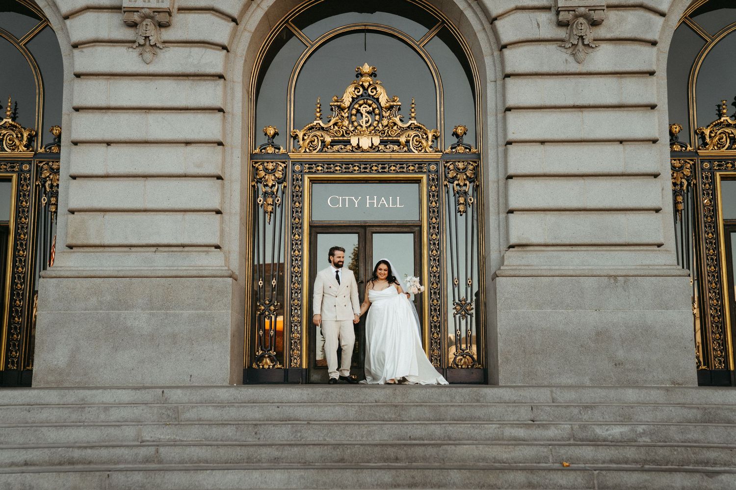 A couple in wedding attire stands in front of an ornate gold-framed mirror at an elegant venue with marble walls.