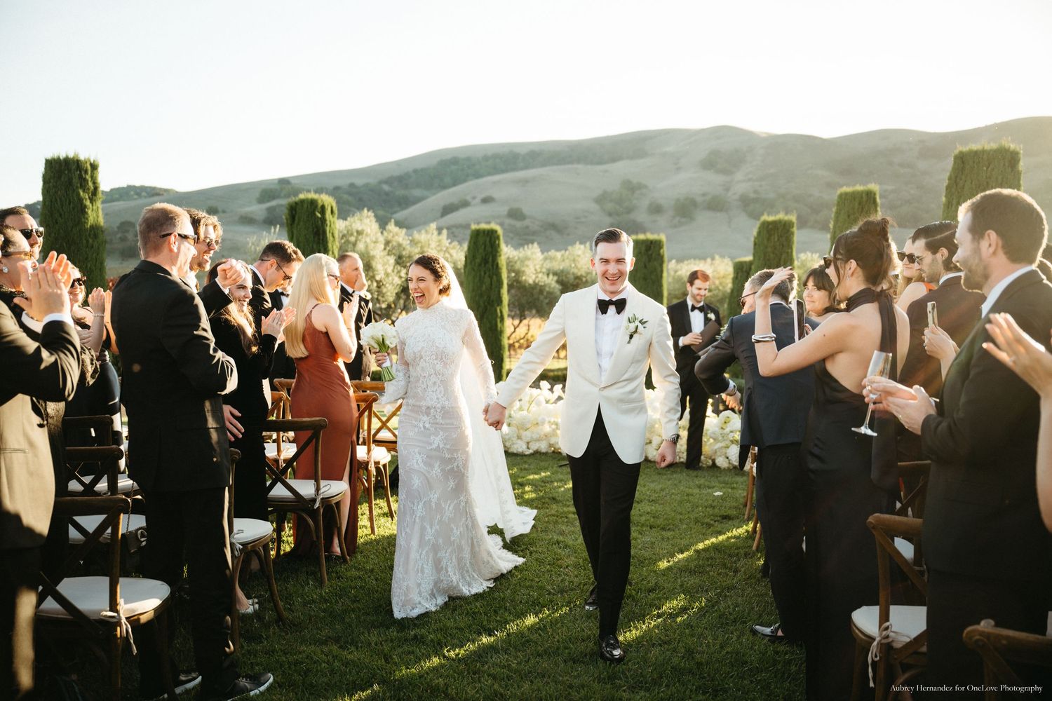 Wedding guests gather outdoors on a grassy hillside during a scenic sunset celebration.