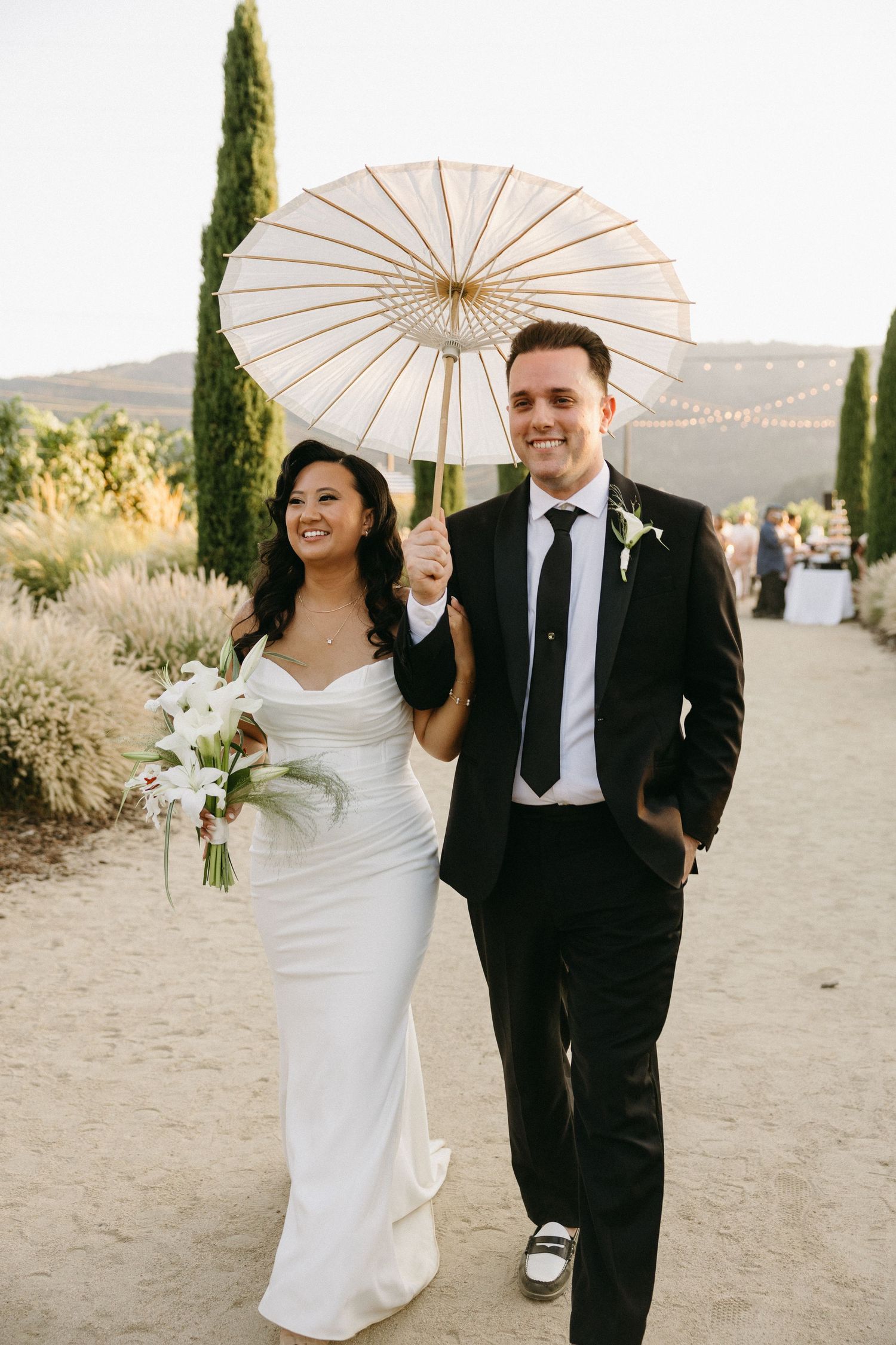 A couple walks together outdoors holding a white parasol at a desert wedding venue during golden hour.