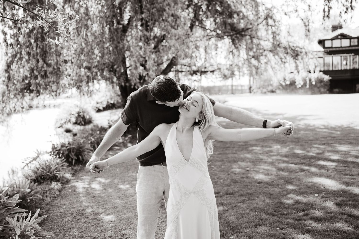 Black and white photo of a joyful couple dancing outdoors near a willow tree, bride in white dress laughing with arms outstretched.