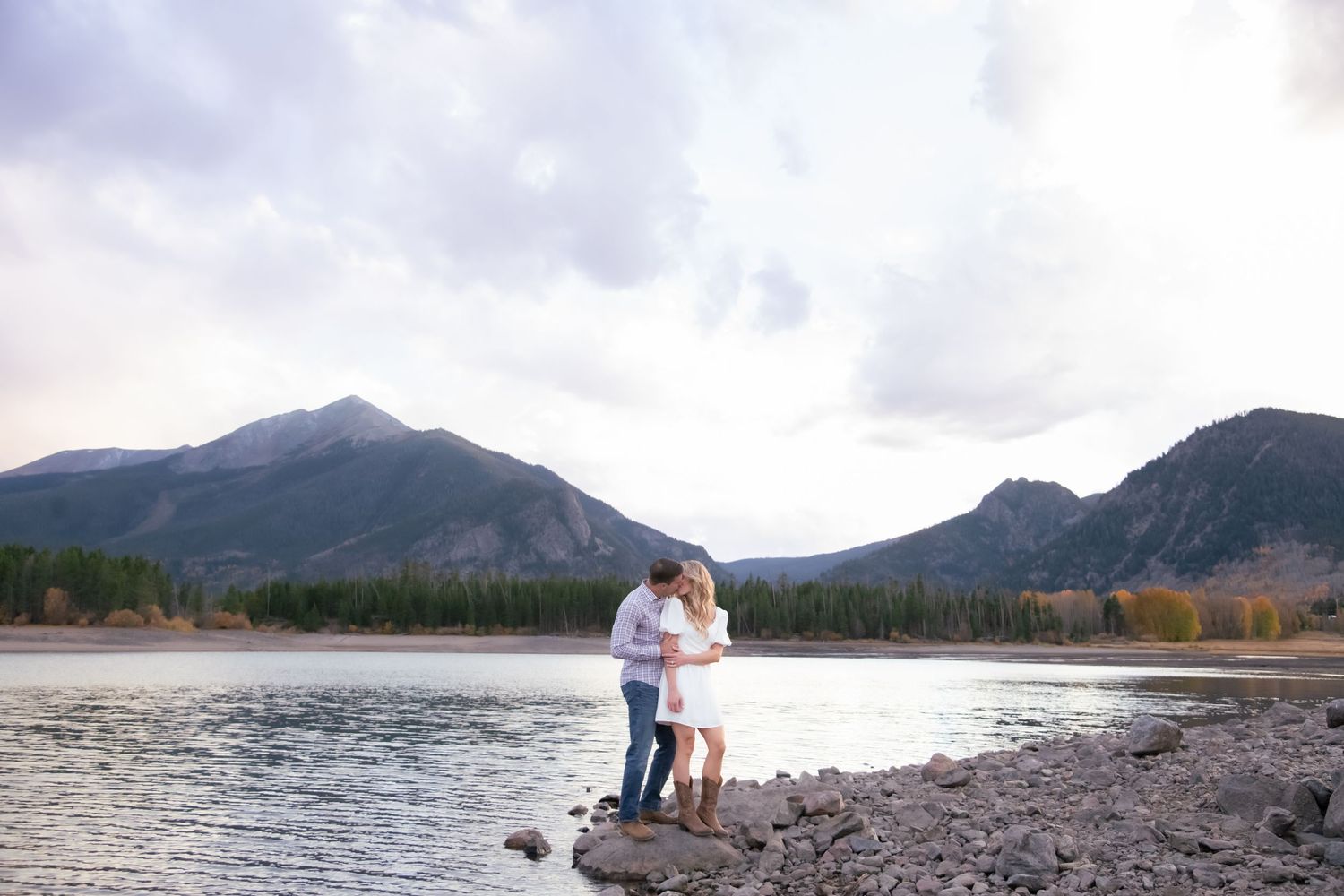 Cute stylish couple stands with water and mountain peaks behind them for an engagement photo session in Breckenridge Colorado
