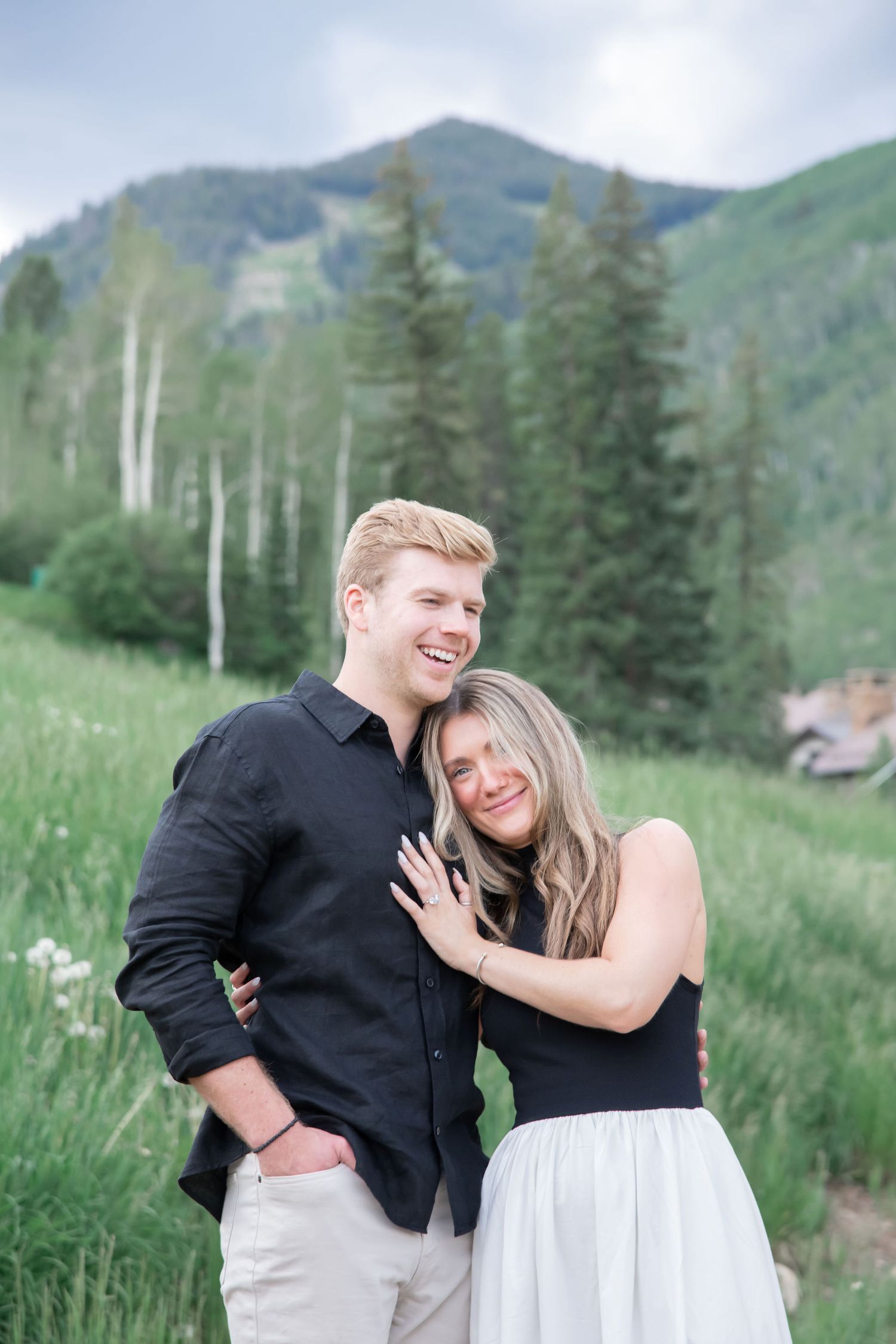 A smiling couple embraces in a lush mountain meadow with mountain in the background for Beaver Creek Colorado engagement.