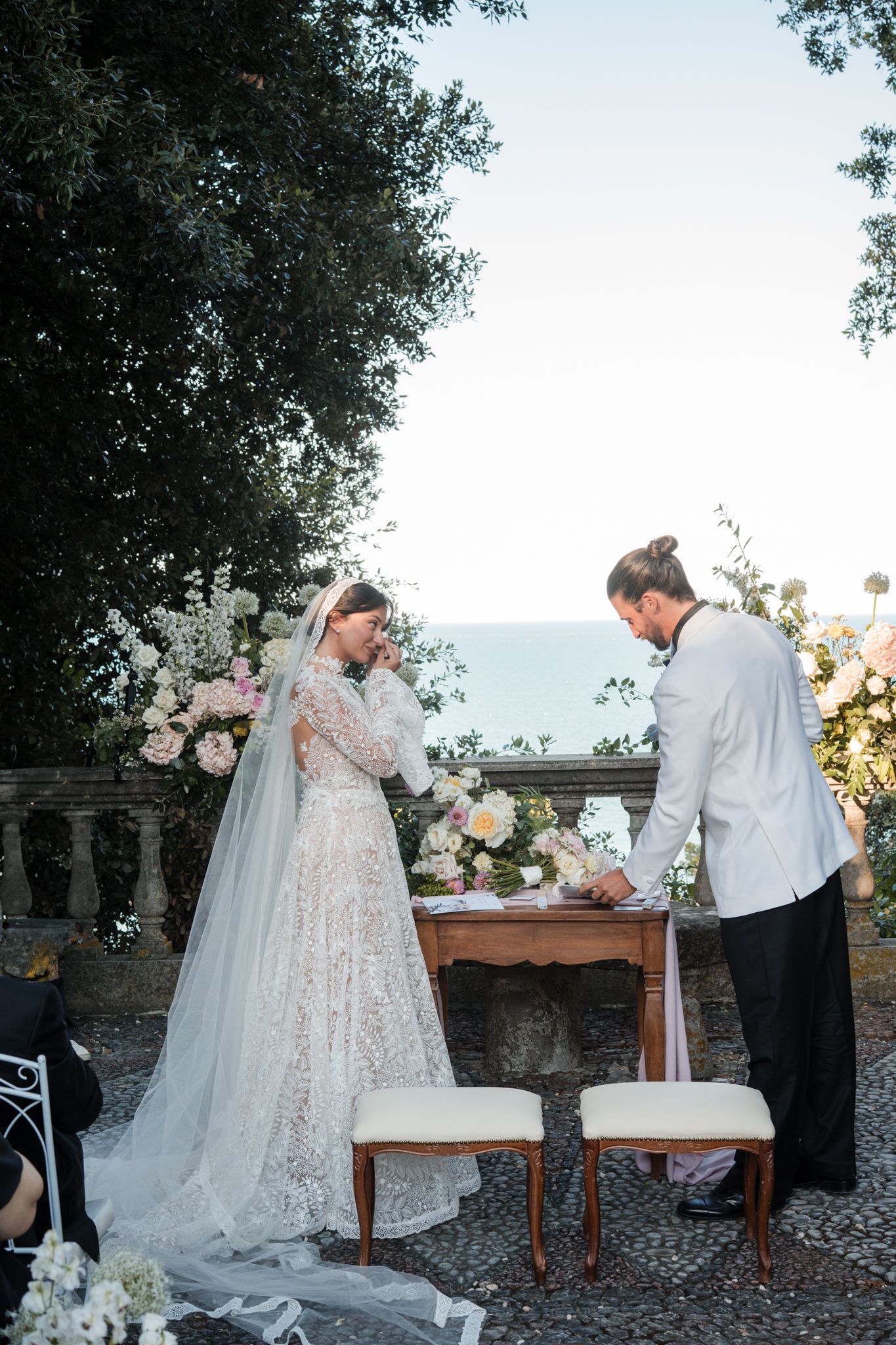 Bride and groom signing wedding register outdoors with floral arrangements and ocean view backdrop.