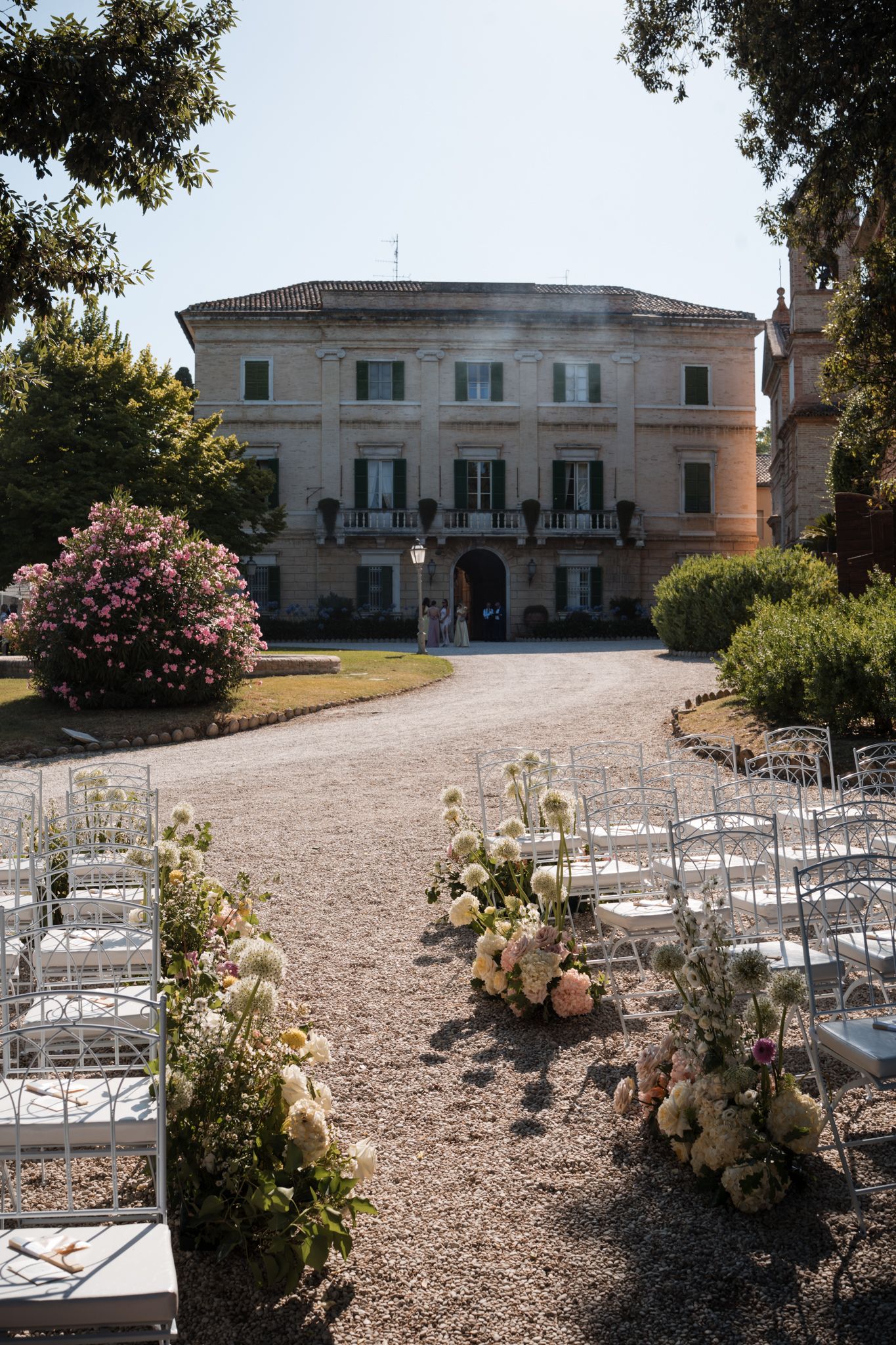 Elegant outdoor wedding ceremony setup with white chairs and floral arrangements leading to a grand Italian villa.