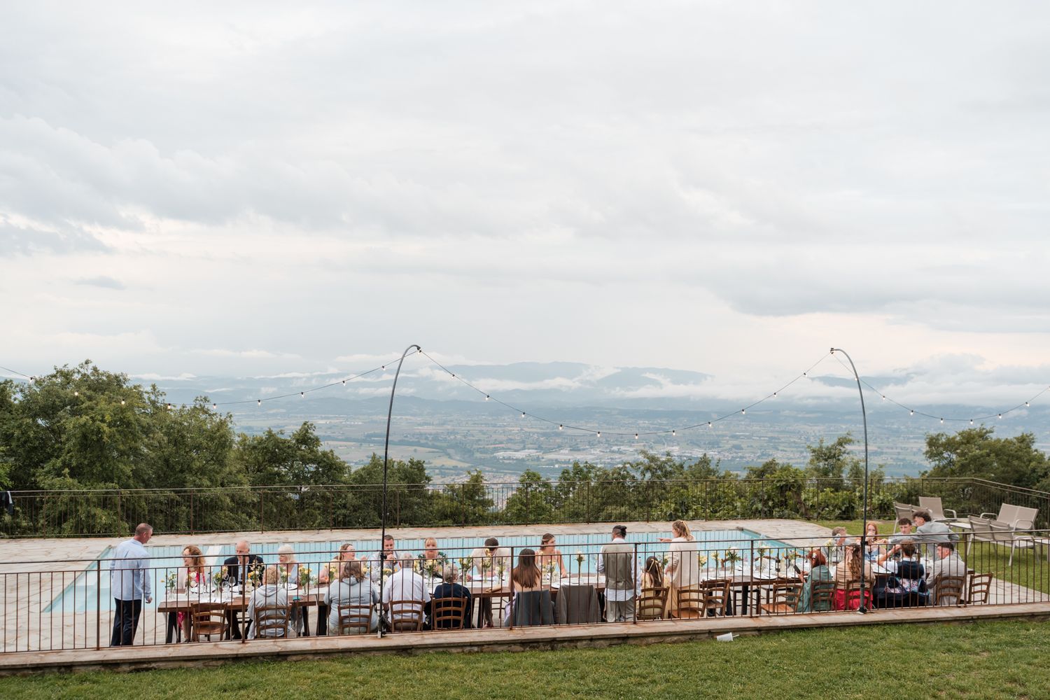Outdoor wedding reception by a pool with panoramic mountain and valley views under a cloudy sky.