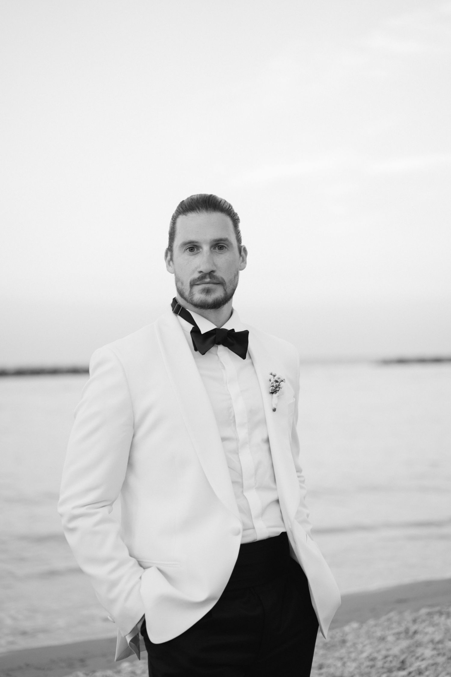 Black and white portrait of a groom in white tuxedo jacket and bow tie standing on a beach.