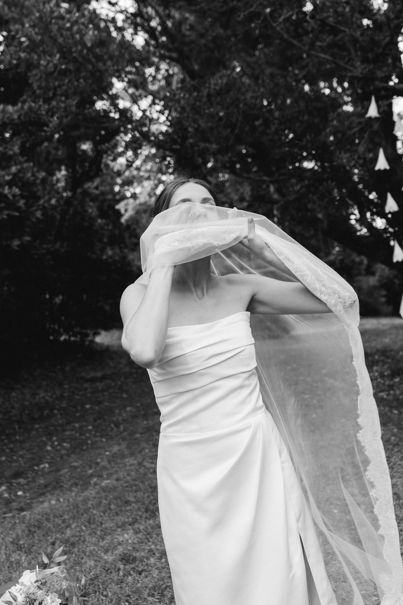 Black and white photo of a bride in a strapless gown holding her lace veil in a lush garden setting.