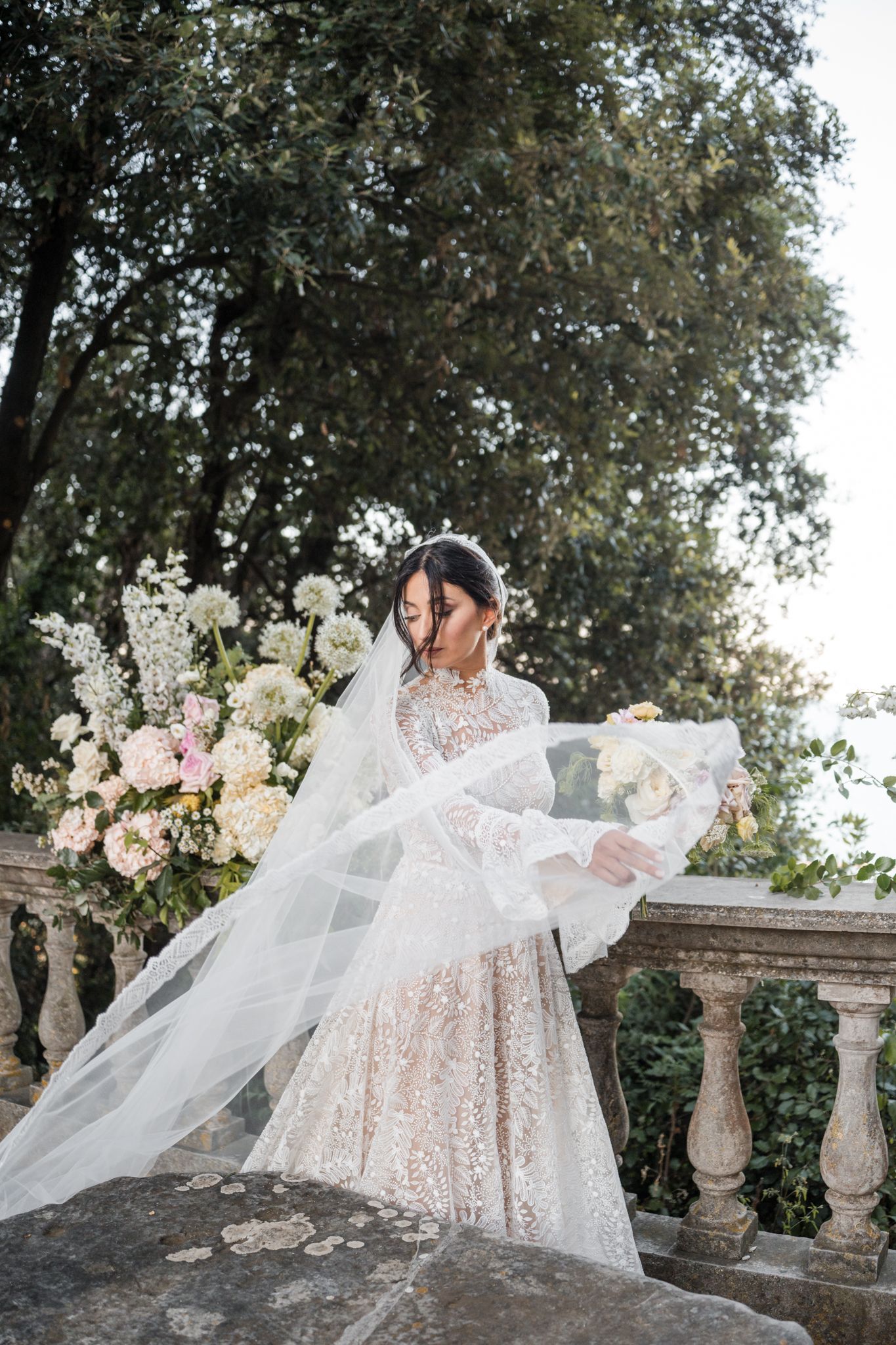 Bride in lace gown with flowing veil stands on stone balcony surrounded by white and pink floral arrangements.