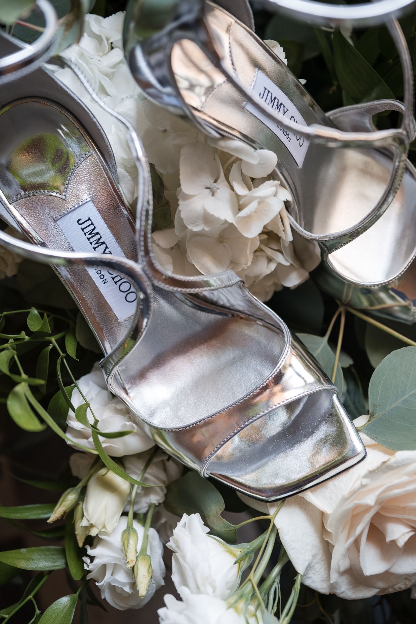 Silver Jimmy Choo strappy heels resting on a bed of white roses and greenery for a bridal flat lay.