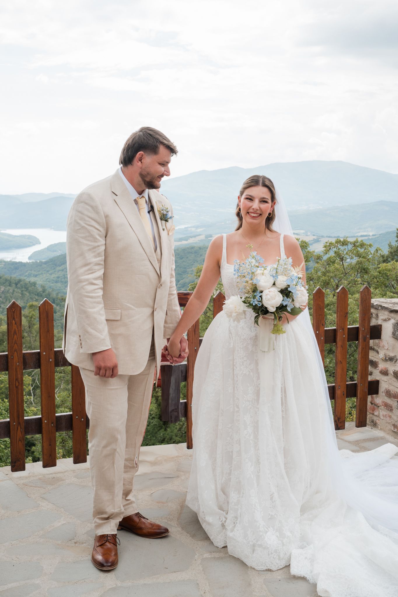 Bride and groom hold hands on a mountain overlook, bride in lace gown with bouquet, groom in beige suit.