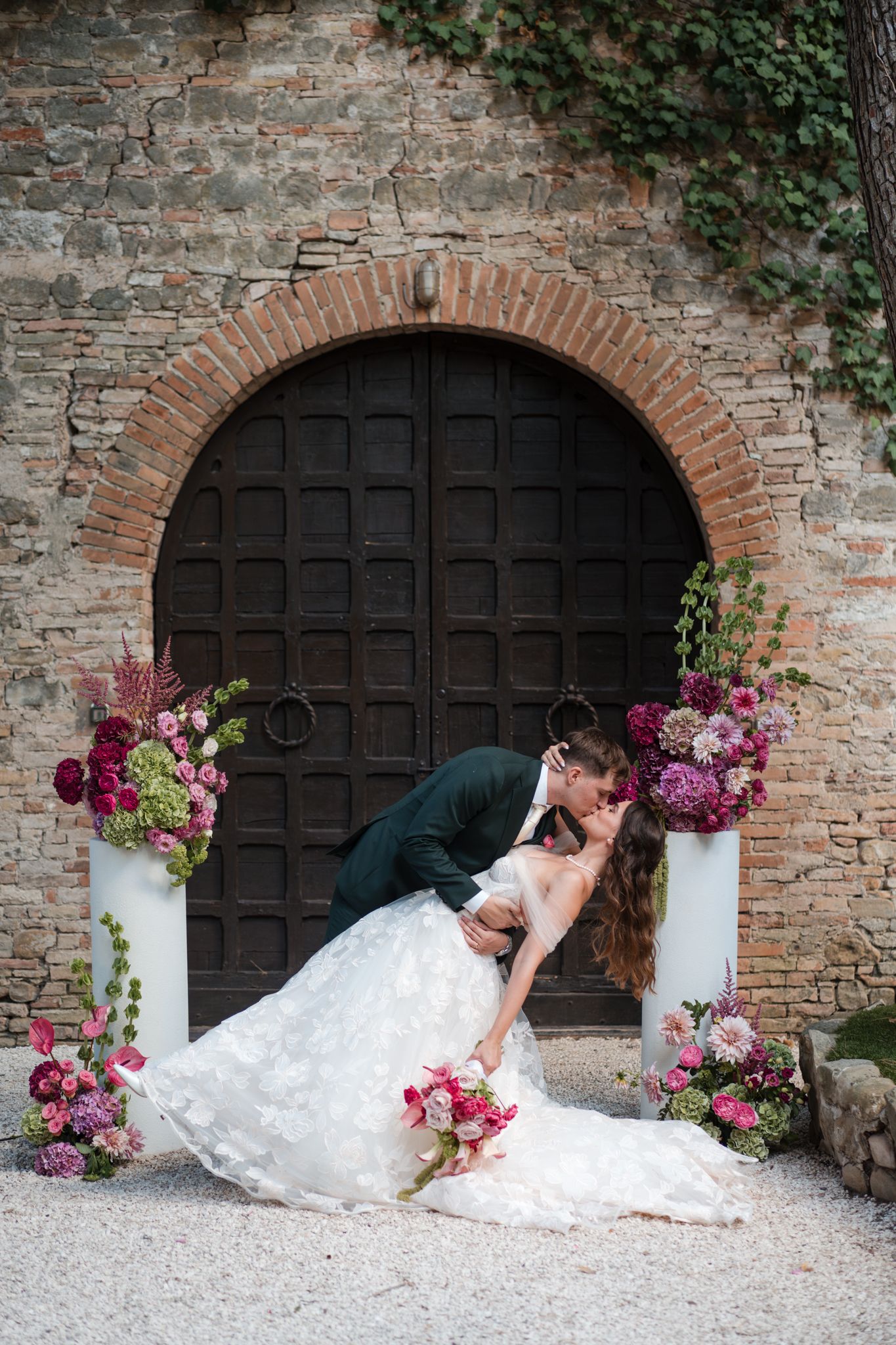 Bride and groom share a romantic dip kiss in front of an arched stone doorway adorned with lush pink and burgundy floral arrangements.