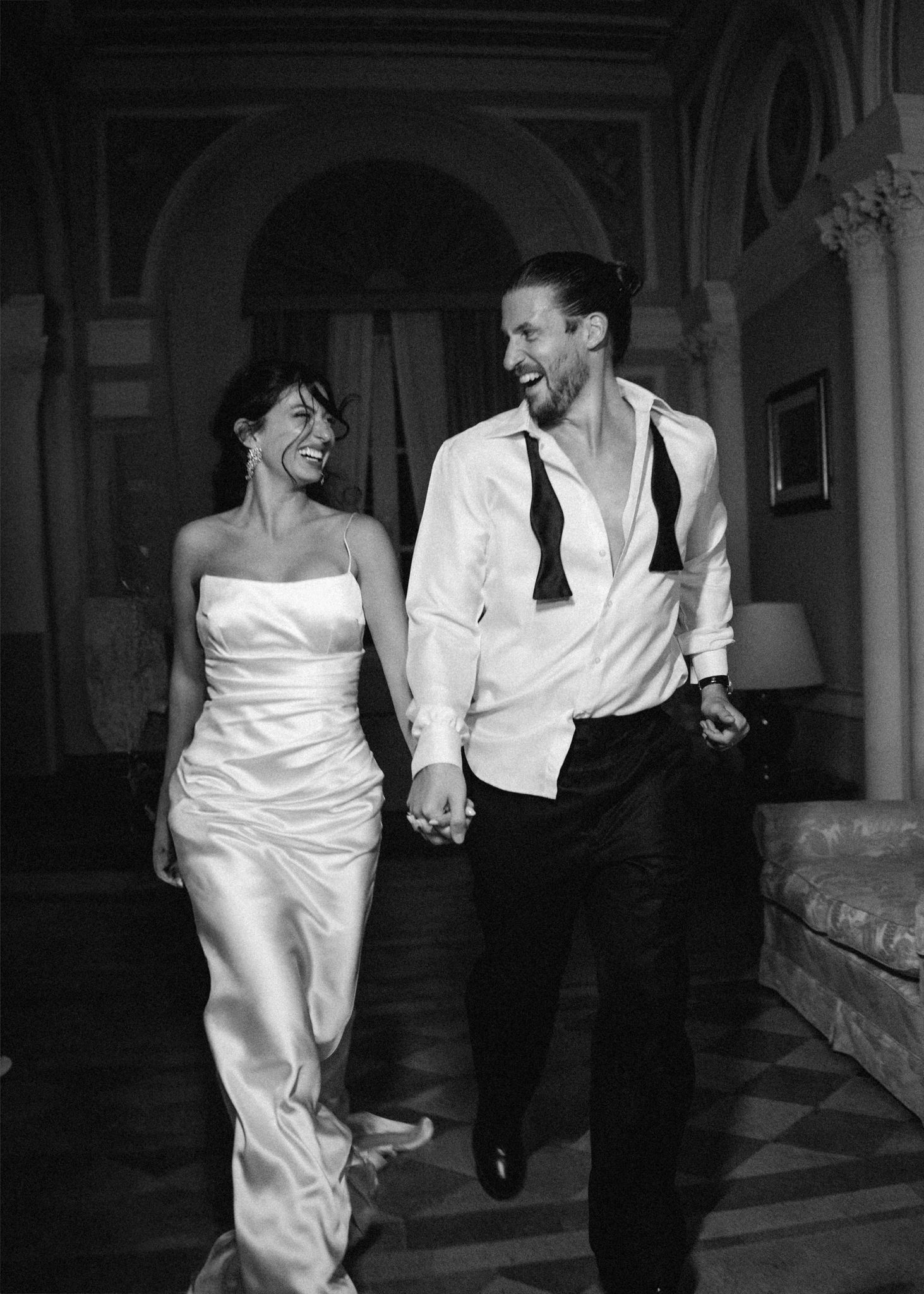 Black and white photo of a joyful couple in wedding attire holding hands and laughing indoors.