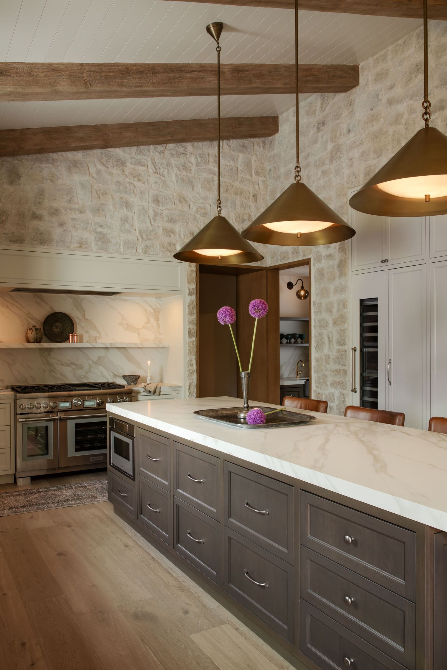 Elegant kitchen with gray island, white marble countertops, brass pendant lights, stone wall, and professional range.