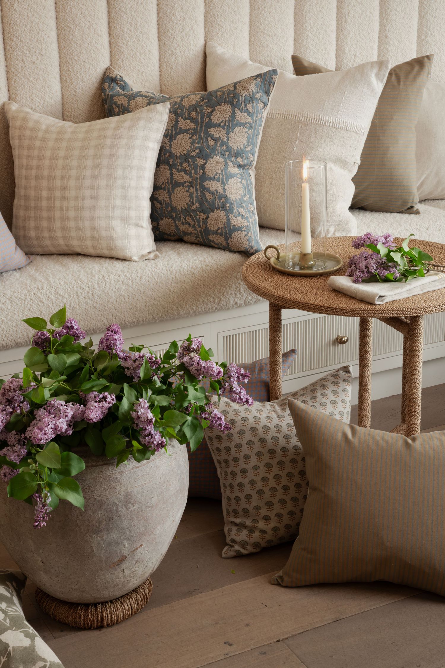 Cozy living room nook with floral and plaid cushions, lilac blooms in stone pot, candle on rattan side table.