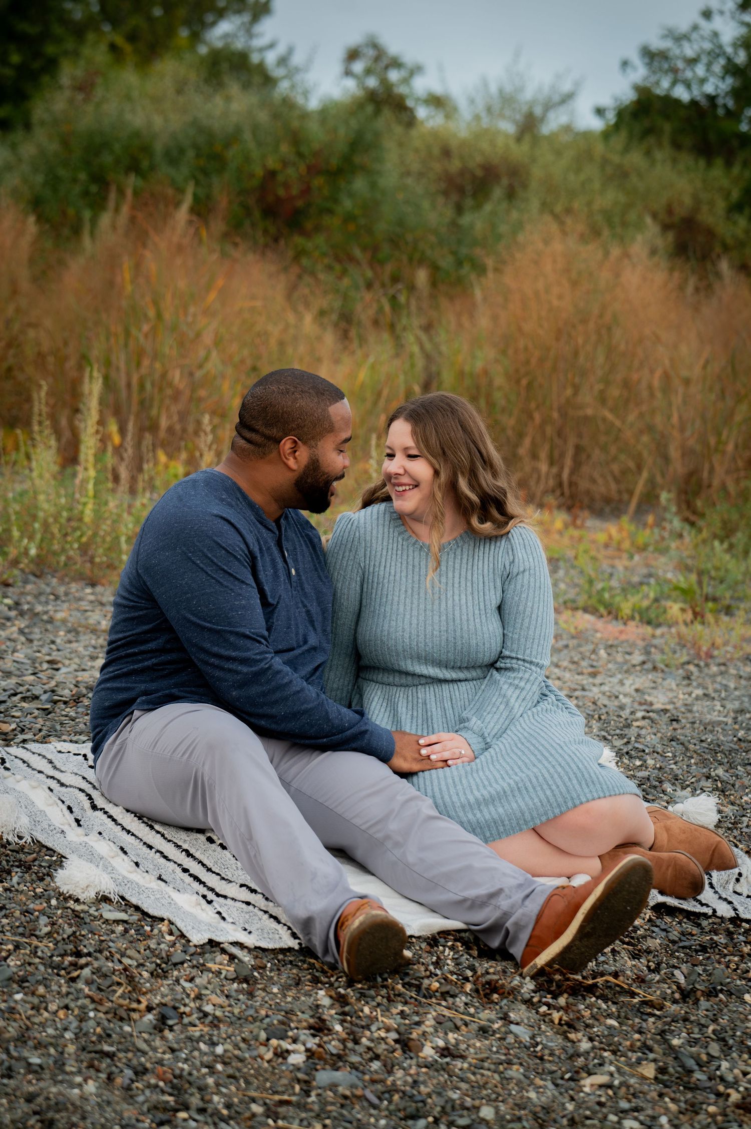 Sweet outdoor engagement photo of couple sitting on blanket sharing tender moment together in rustic setting at sunset.