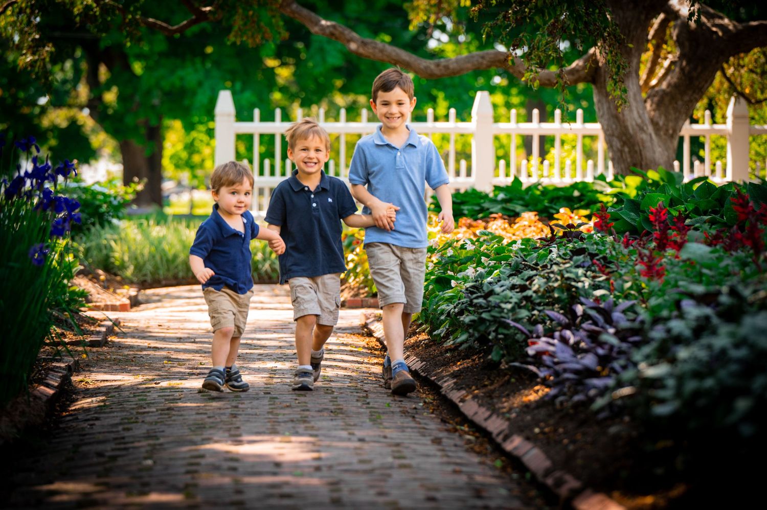 Three siblings walk together along a garden path lined with colorful flowers and a white picket fence on a sunny day.