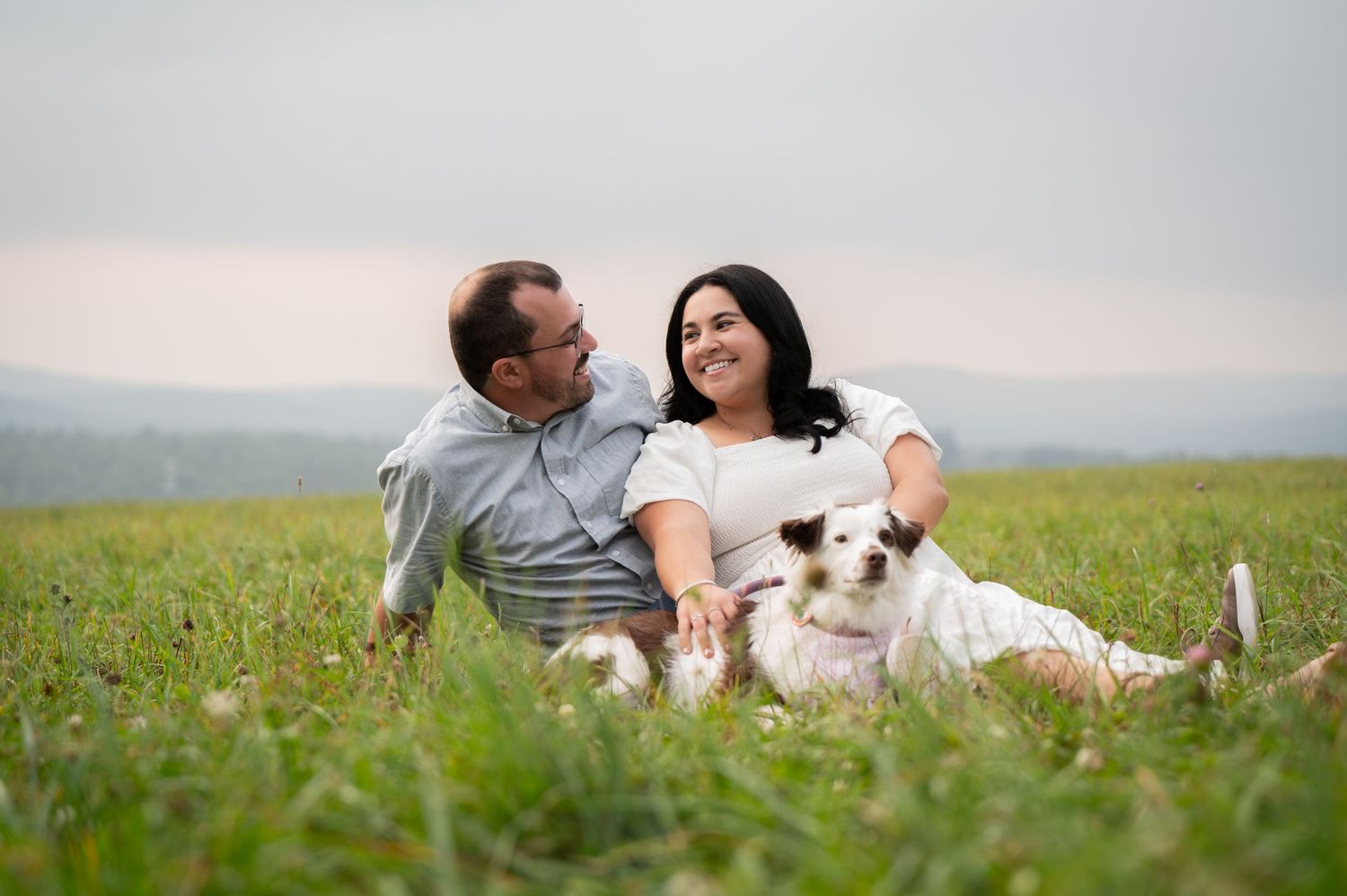 Couple relaxing in grassy meadow with their white dog on a cloudy afternoon.