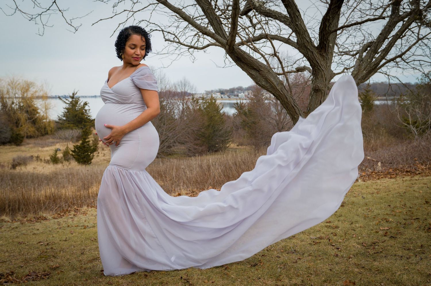 A flowing white maternity dress billows dramatically in the wind against a natural outdoor backdrop of bare trees and dry grass.