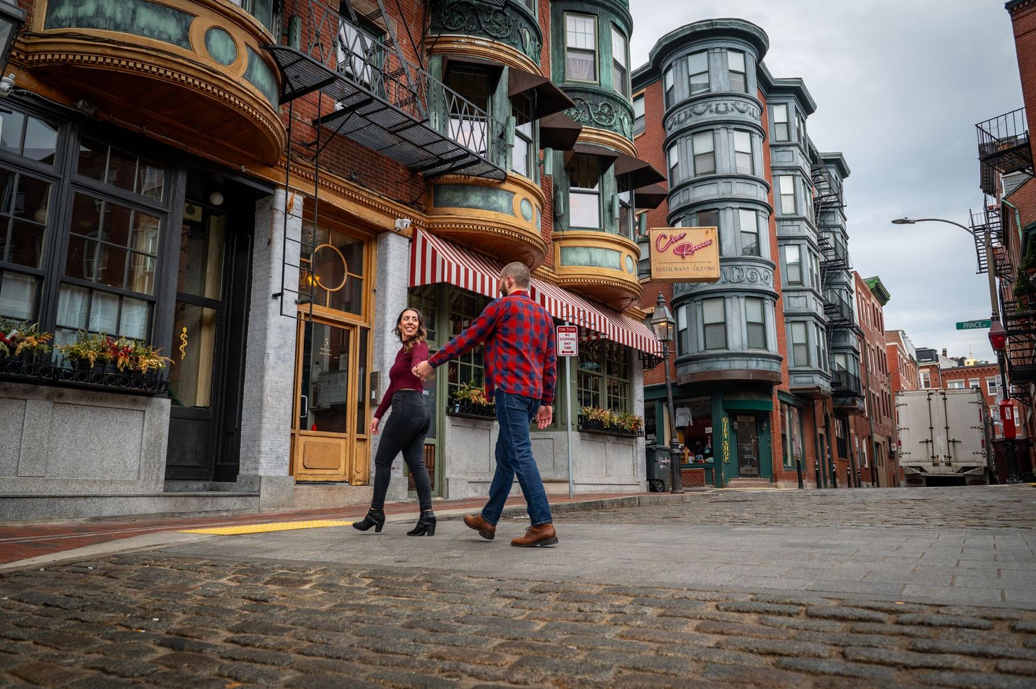 People walk past historic Victorian buildings on a cobblestone street in Boston's North End neighborhood.