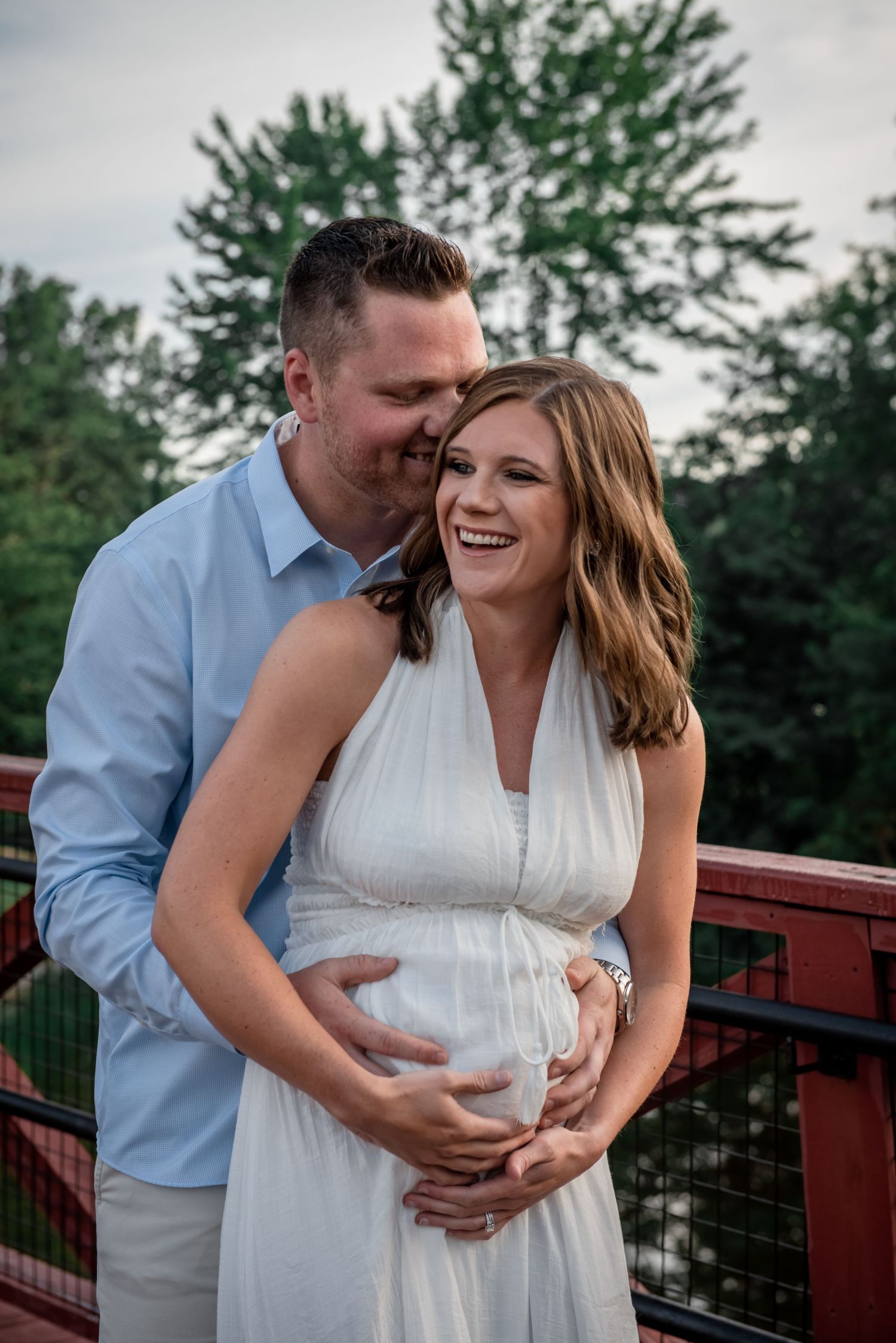 A joyful expecting couple embraces on a red bridge outdoors during a maternity photo shoot.