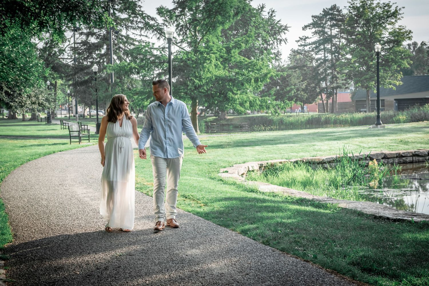 A couple takes a romantic stroll along a winding path through a lush green park on a misty summer day.