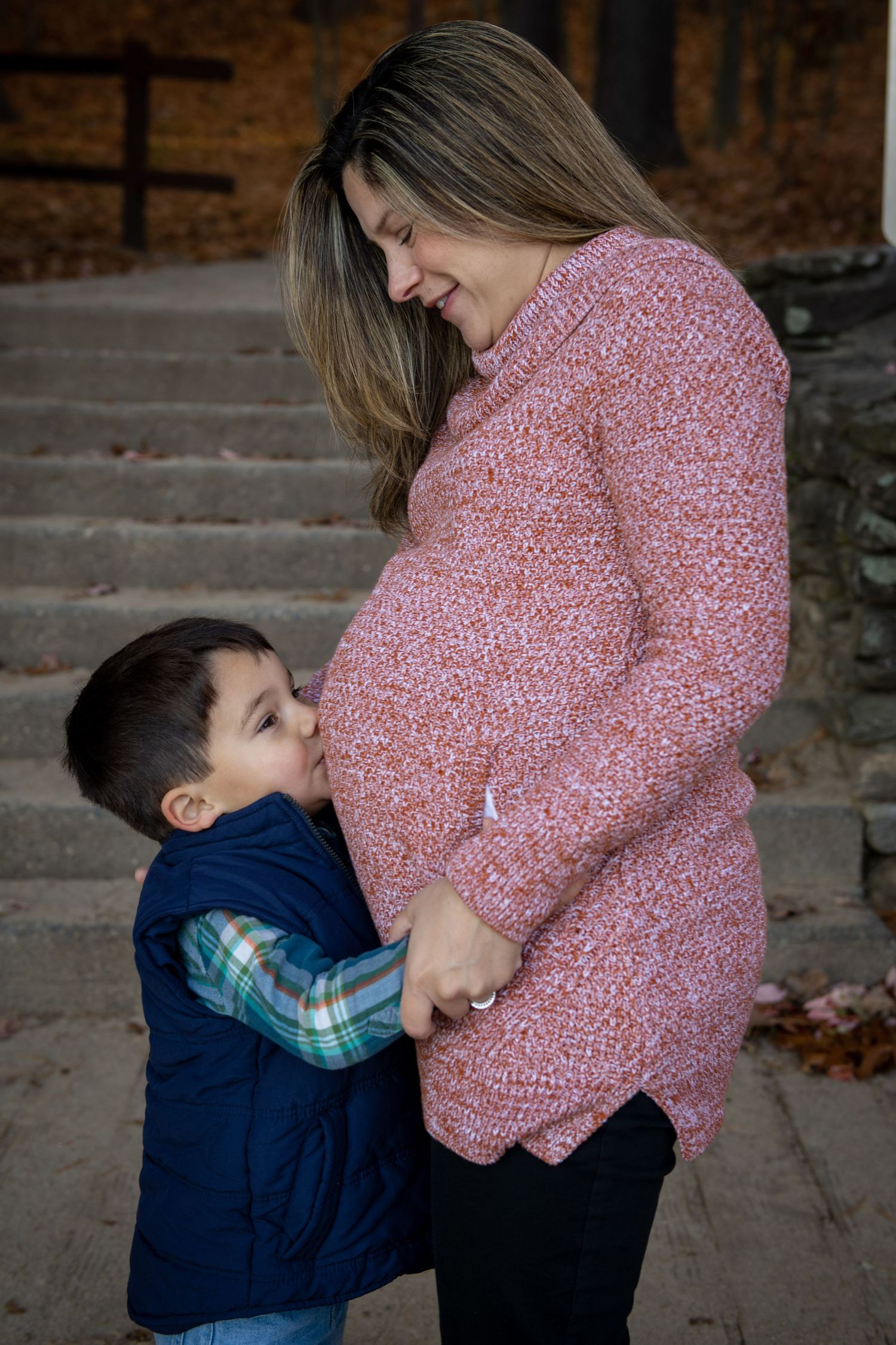 A young child hugs a pregnant woman's belly on stone steps during an autumn day while wearing a navy vest.