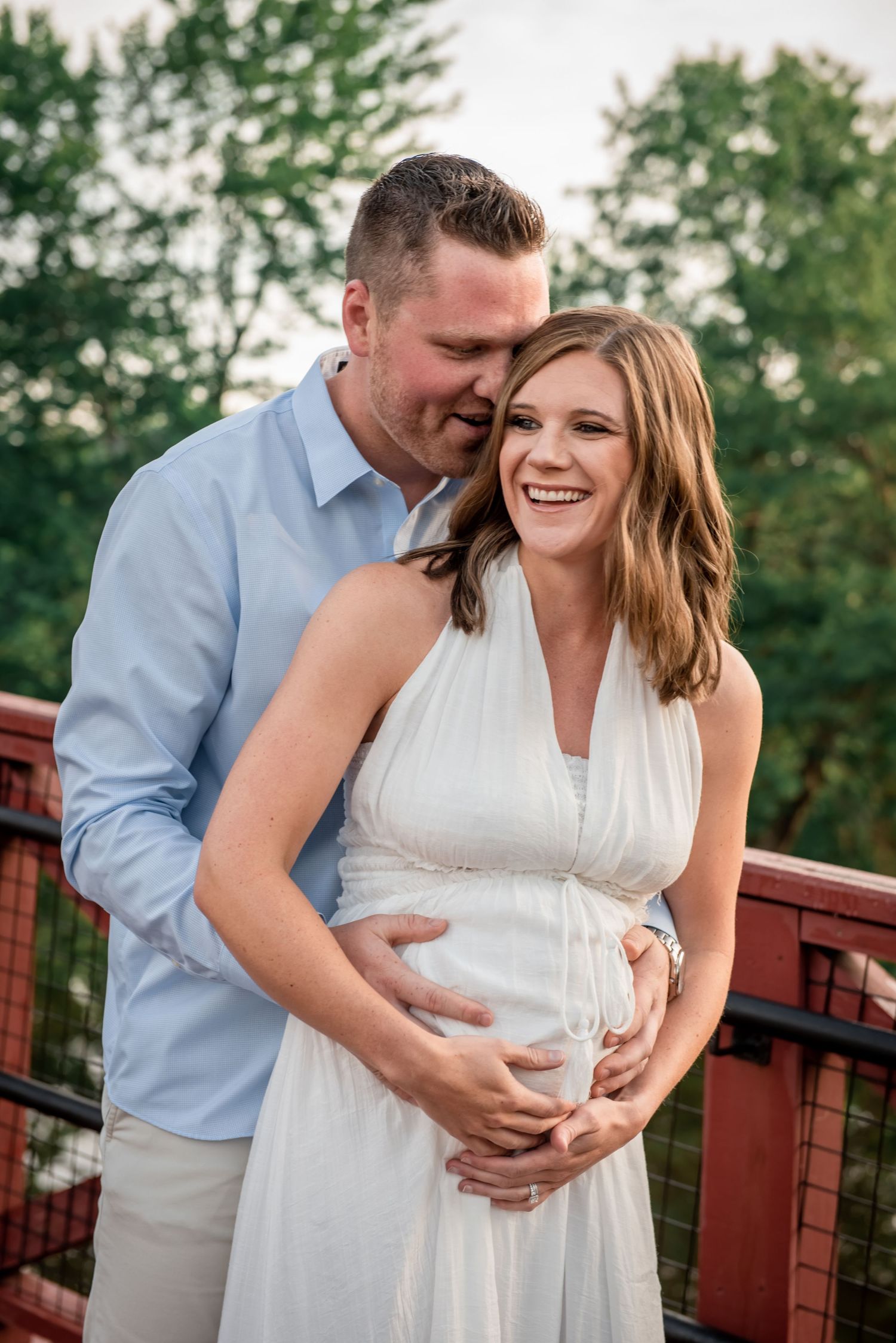 A loving couple shares an intimate moment on a deck during a maternity photoshoot, with green trees in the background.