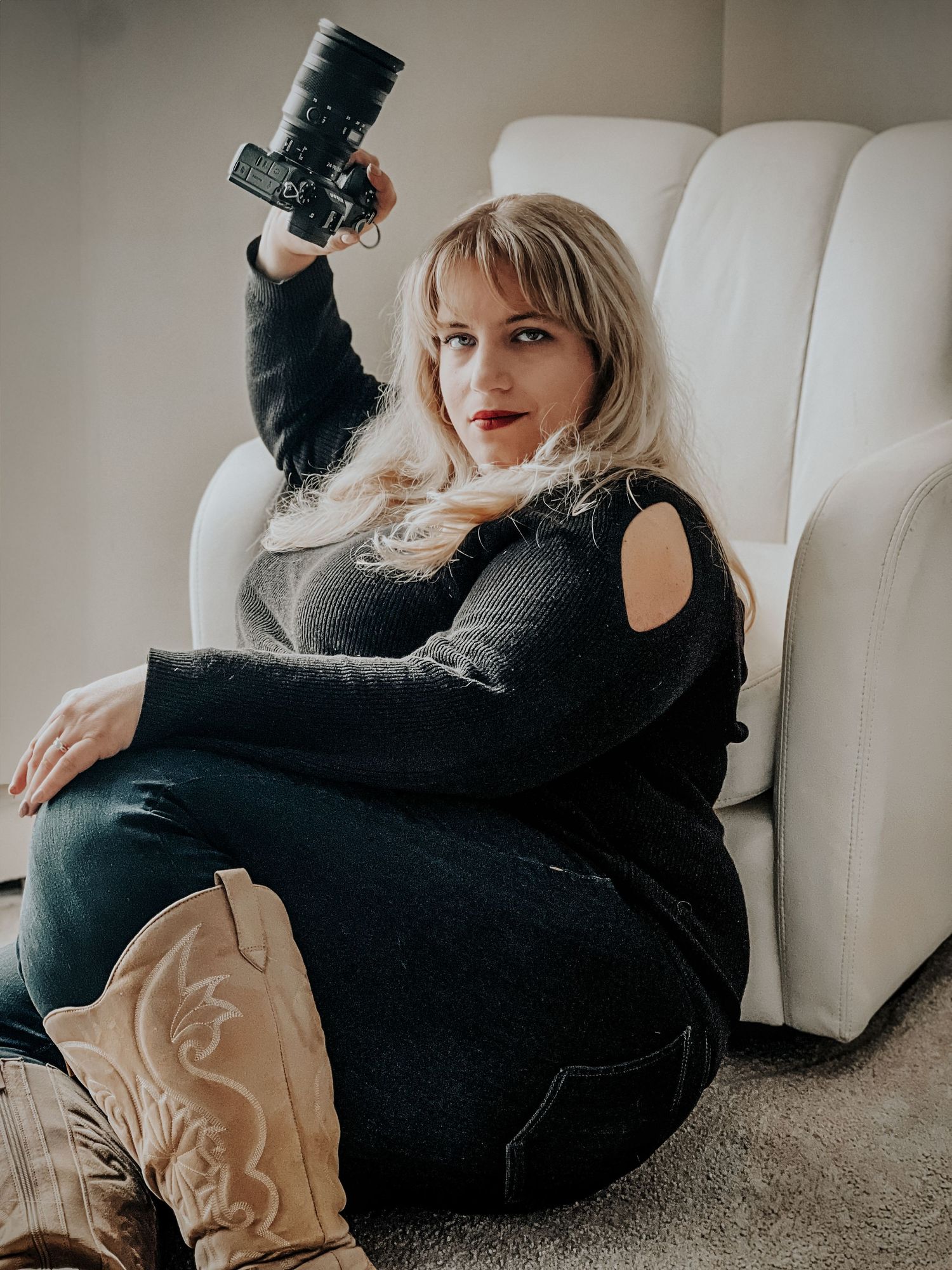 A photographer relaxes on a white couch holding a camera while wearing black clothing and tan cowboy boots.