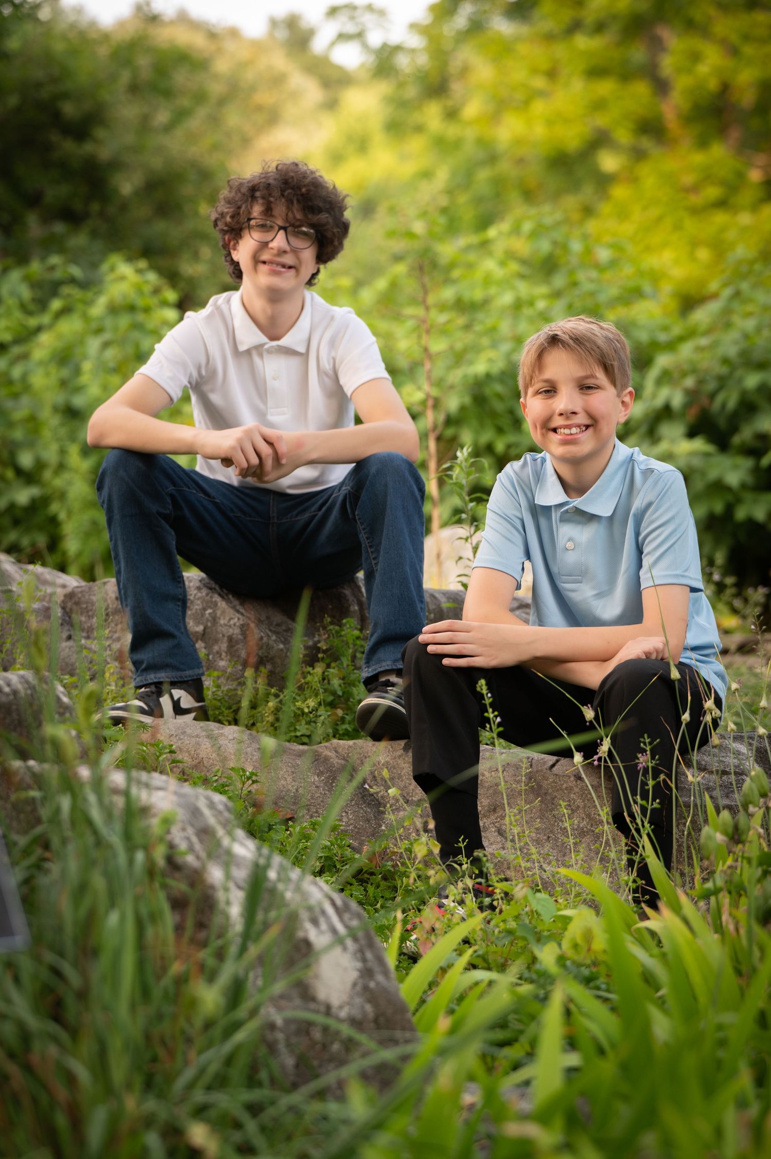 Two boys in casual attire sit together on large rocks surrounded by lush greenery on a sunny afternoon.