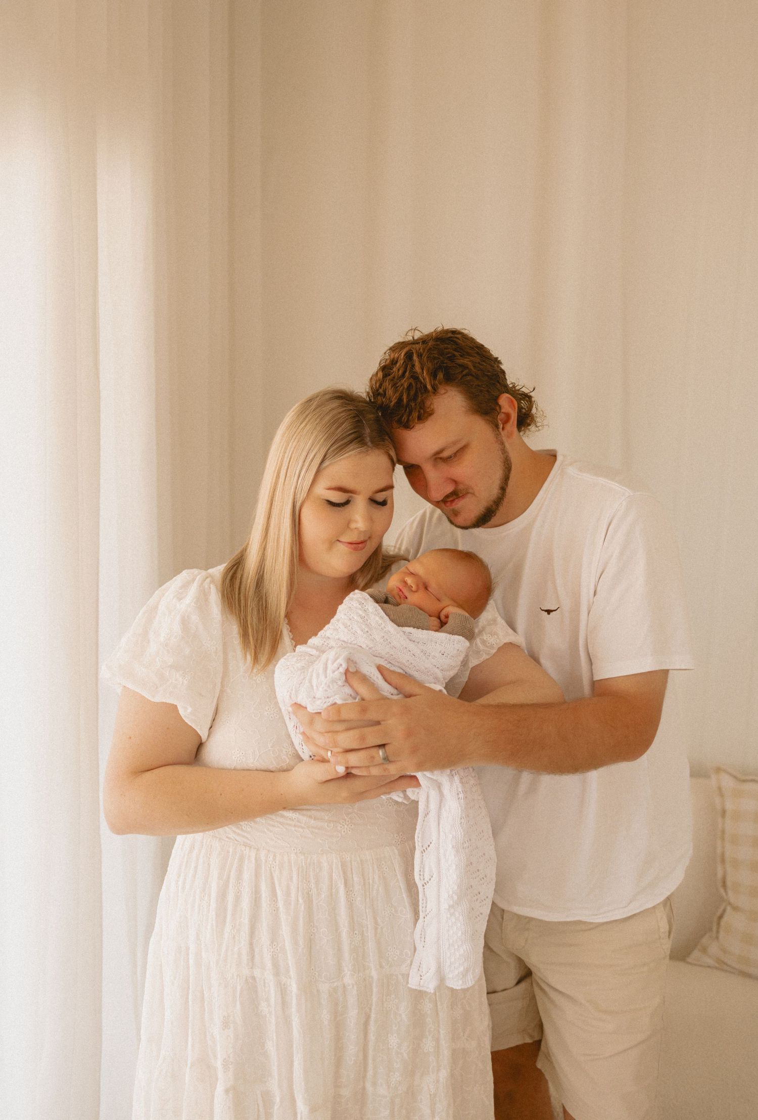 Couple in white clothing tenderly holding their newborn baby against a light curtain backdrop.