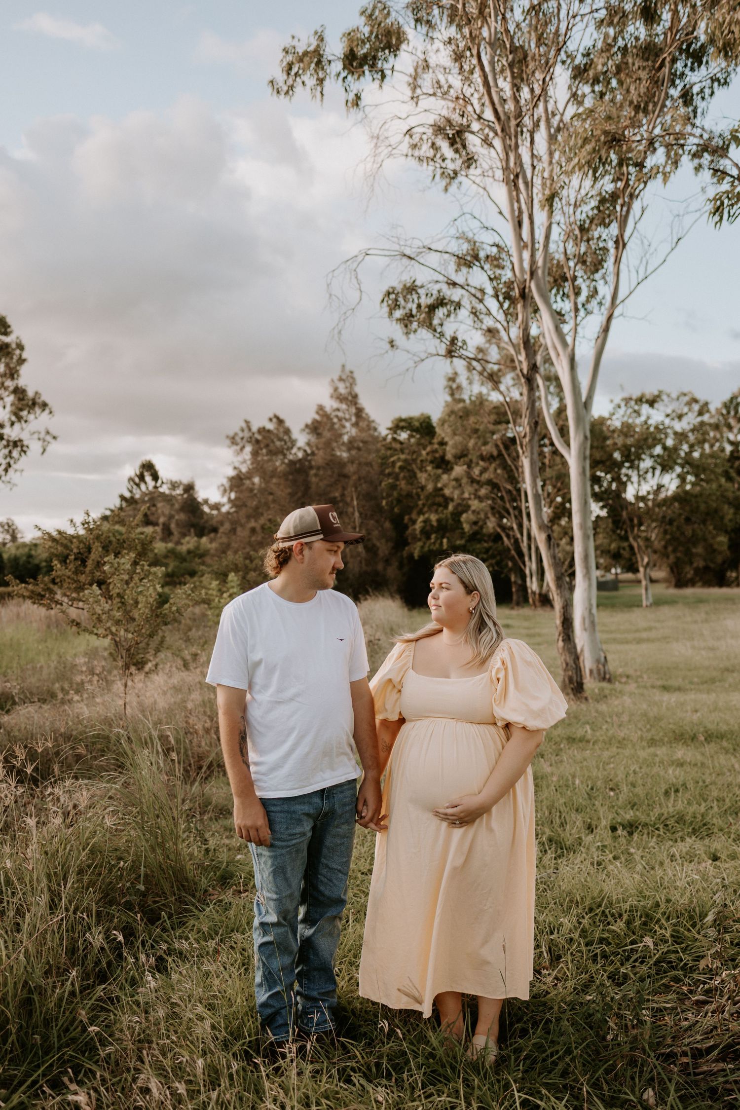 Couple stands together in golden field with tall eucalyptus trees during sunset maternity photoshoot.