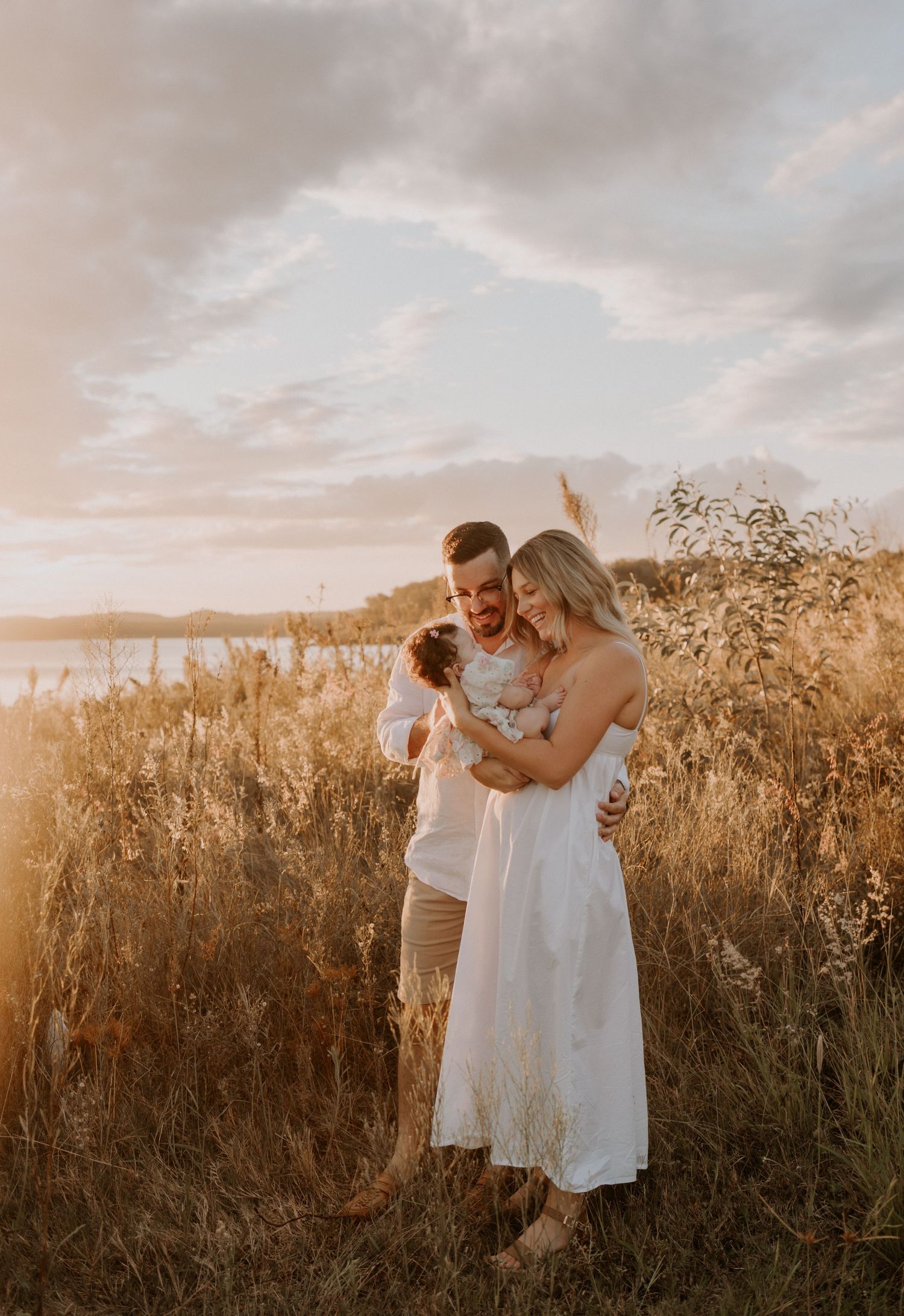A couple embraces with their baby at sunset in a golden field of tall grass with water in the background.