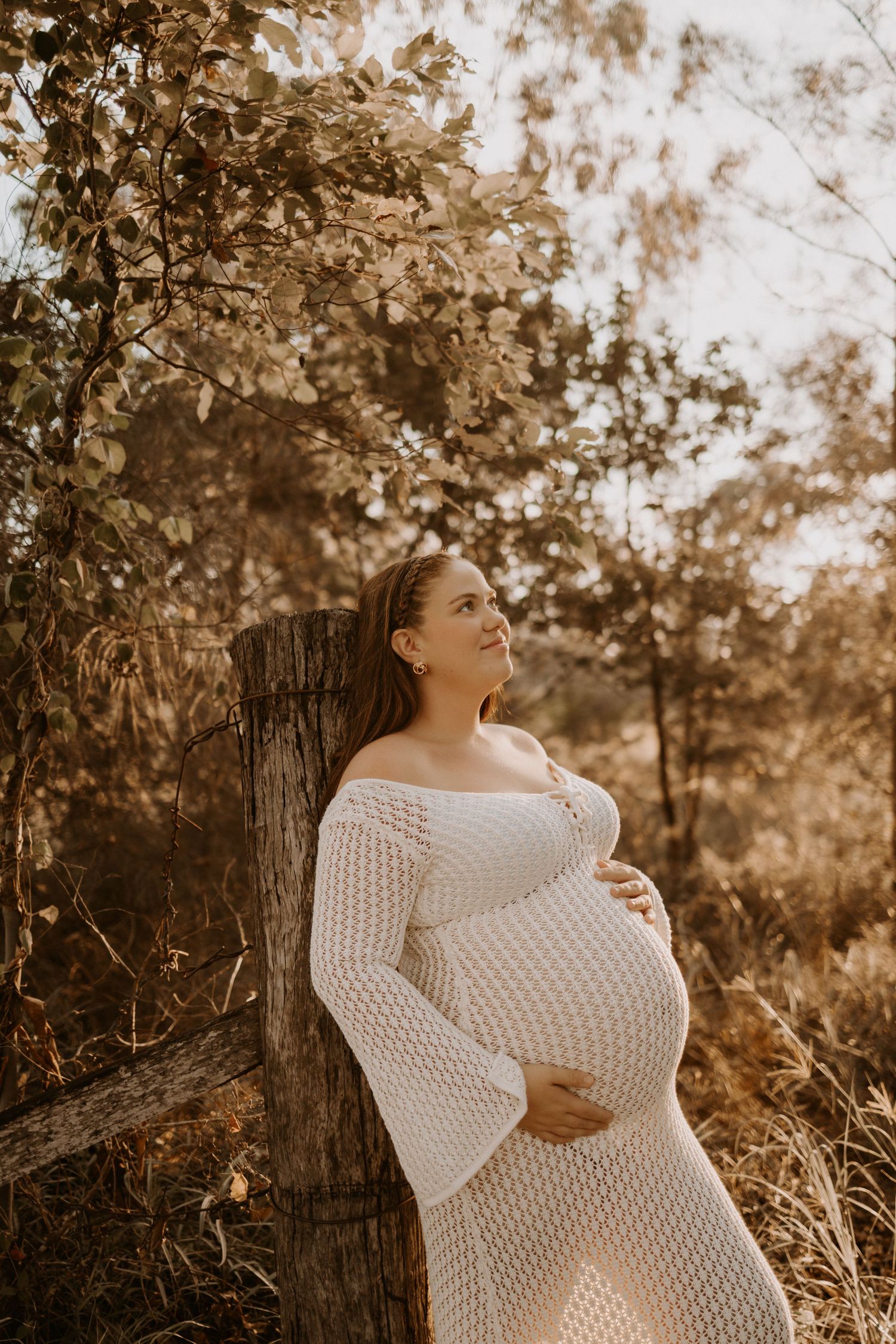 A pregnant woman in a white knit dress poses outdoors in warm sepia tones against a rustic wooden fence post.
